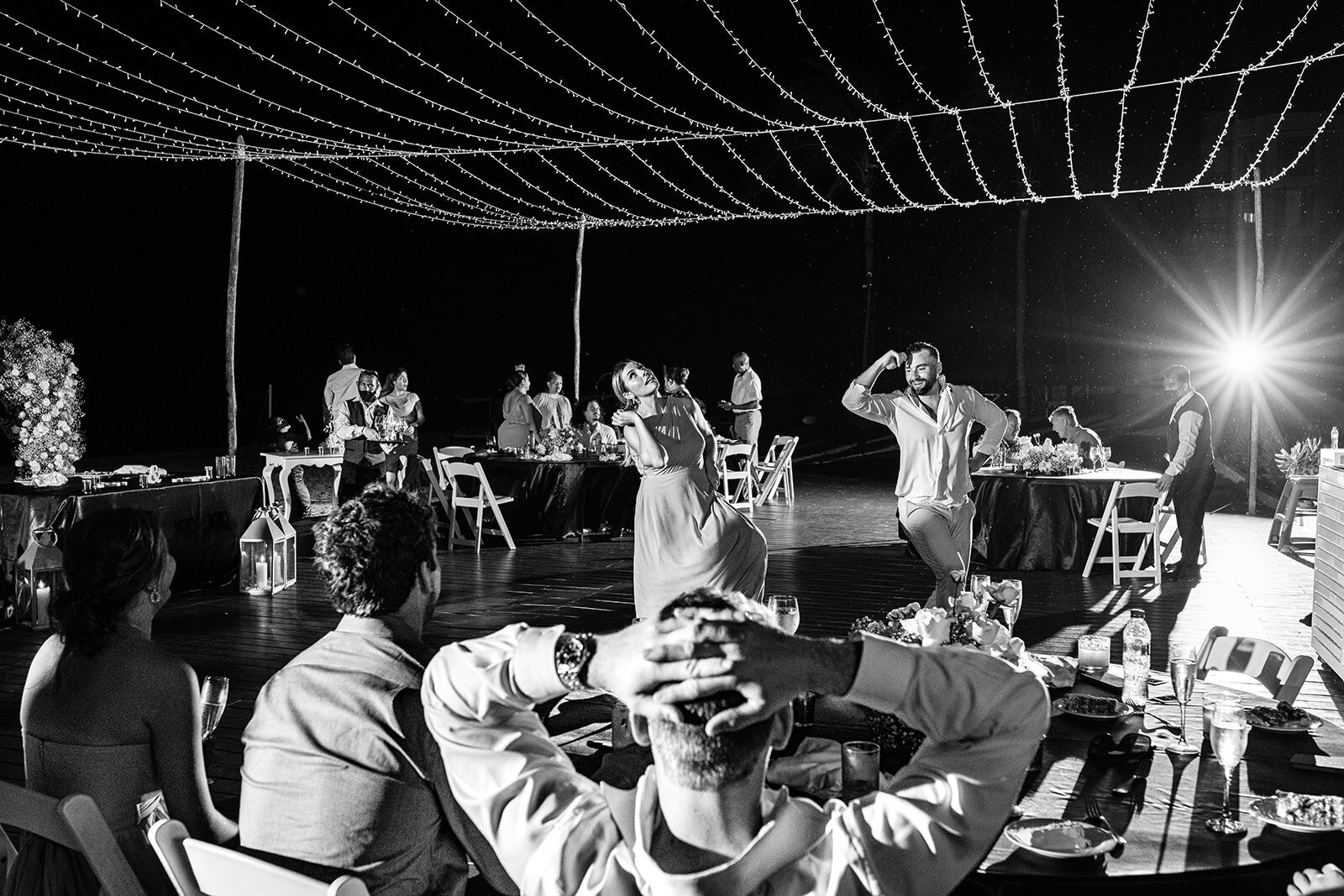 A fun and candid moment of guests dancing and enjoying themselves under a canopy of string lights during an outdoor wedding reception, captured in black and white by CMH Studios