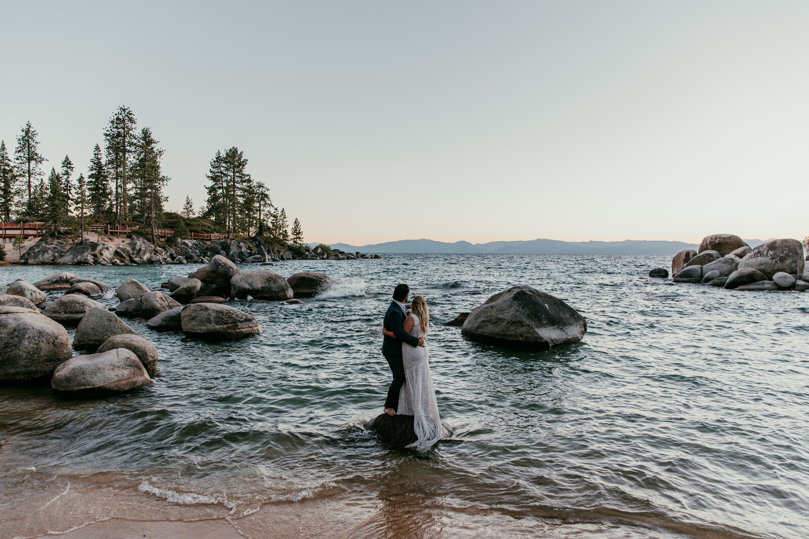 Couple standing barefoot on a rock at the edge of Lake Tahoe during their sunset elopement.