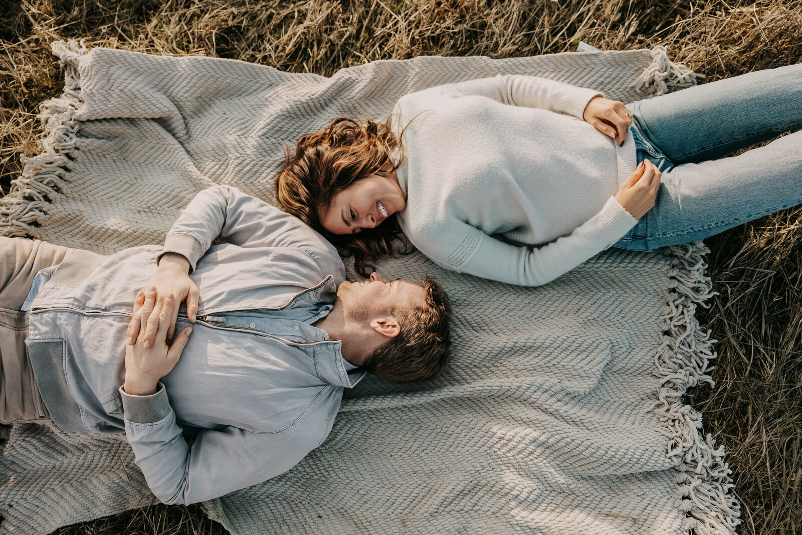 A couple lying on a blanket in a field, heads together, smiling at each other under the warm sunlight.
