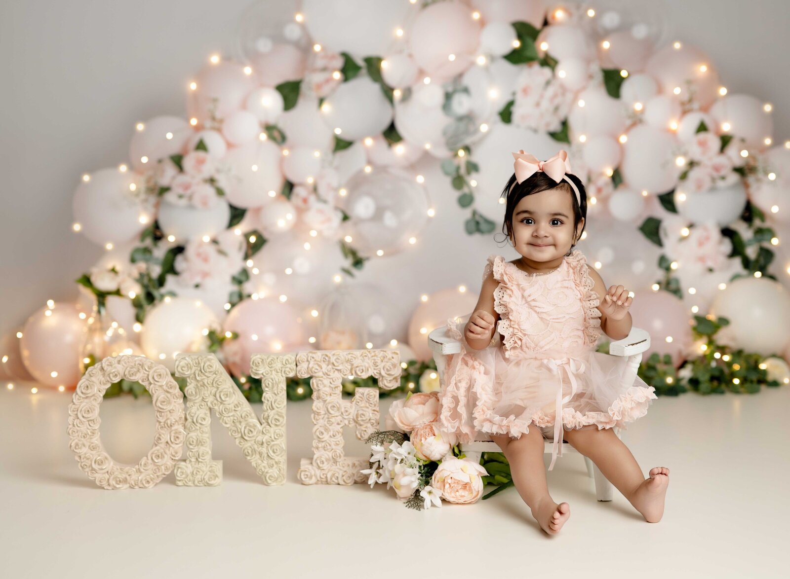 Baby girl taking 1st birthday photo, sitting in a chair next to large ONE letters. 1st birthday photo taken by Jenni Nikki Photography in Atlanta, GA.