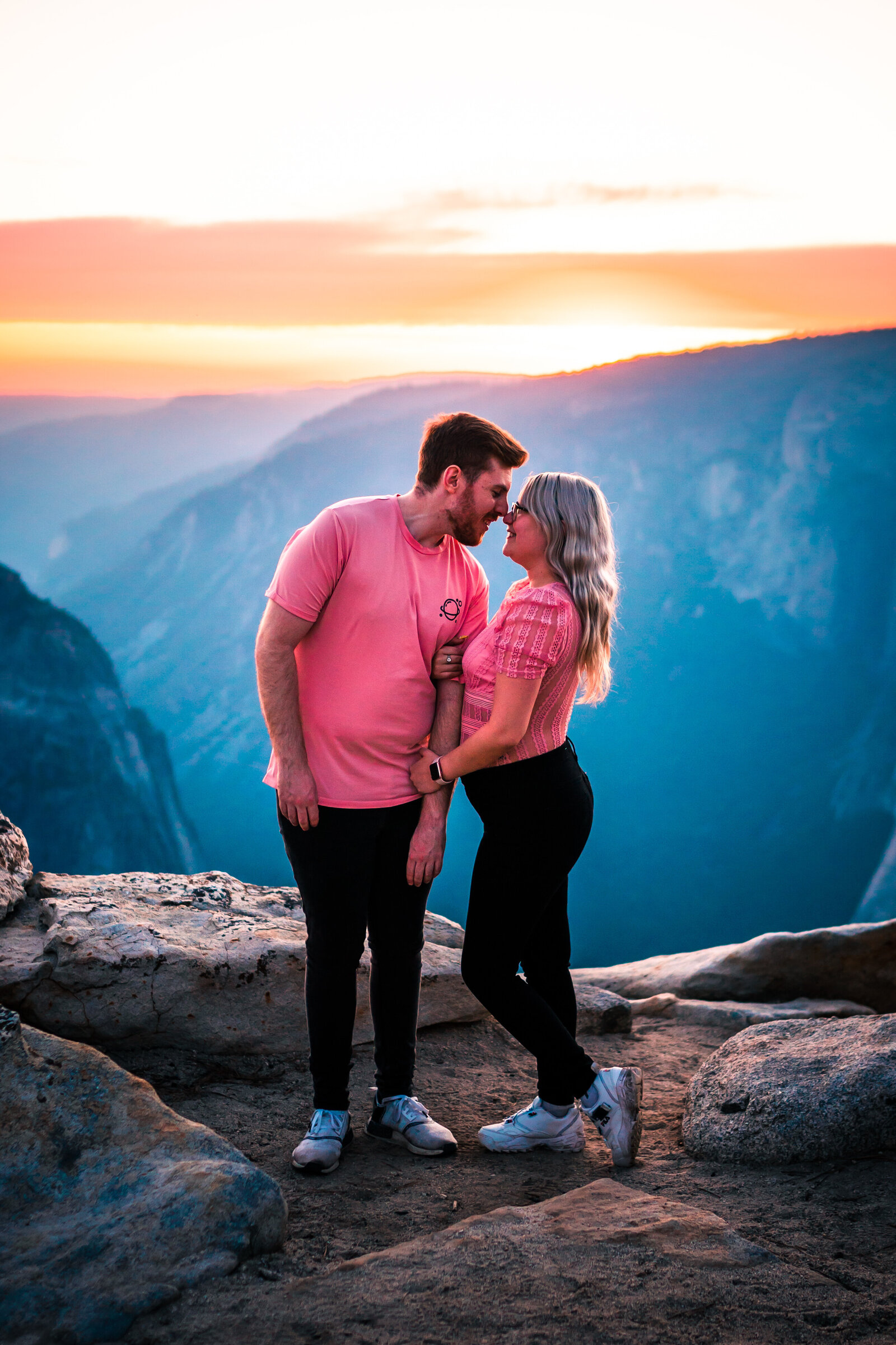 Couple wearing matching clothes, looking at each other with noses touching and smiling, with mountains and beautiful sunset in background.
