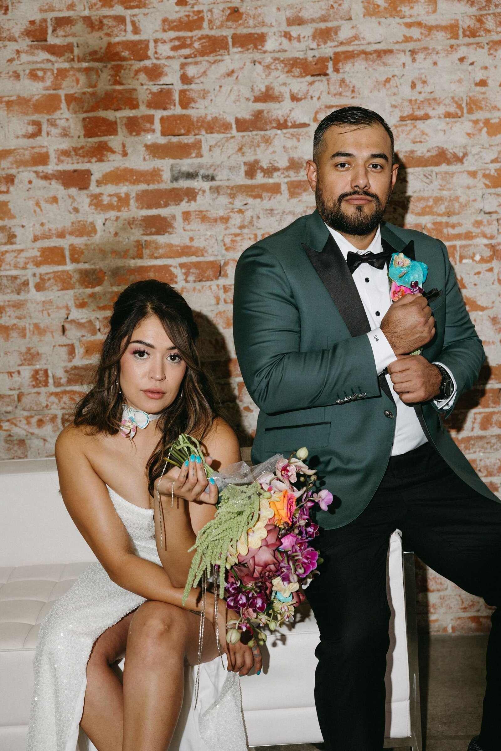 A bride and groom sit on a white sofa, the bride holds a colorful bouquet and the groom adjusts his sleeve