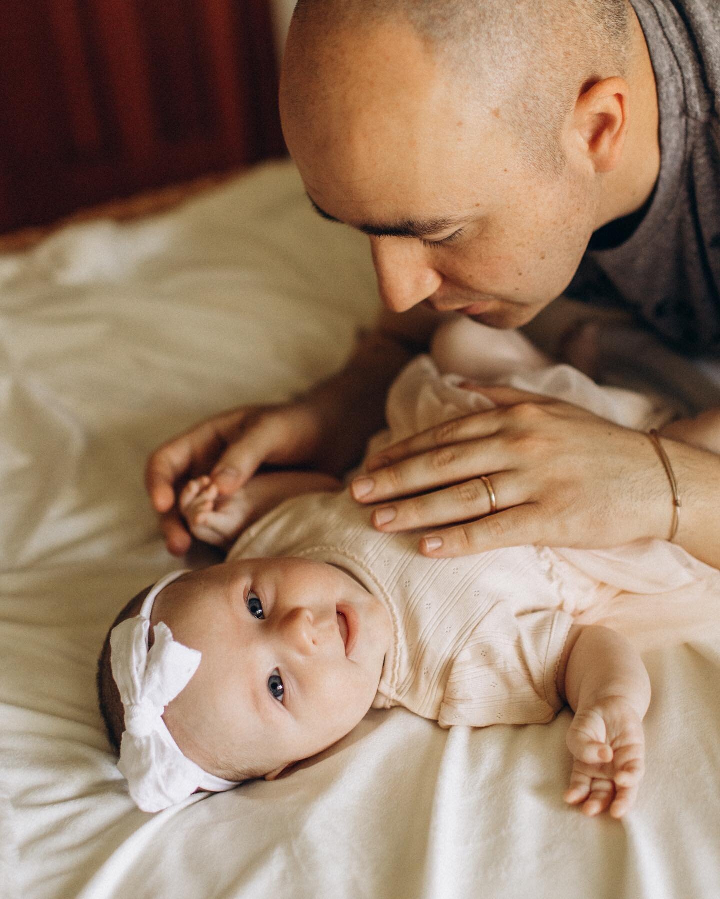 A bald man gently touches a smiling baby lying on a bed, both looking at each other. the baby wears a white bow headband and a cream onesie.