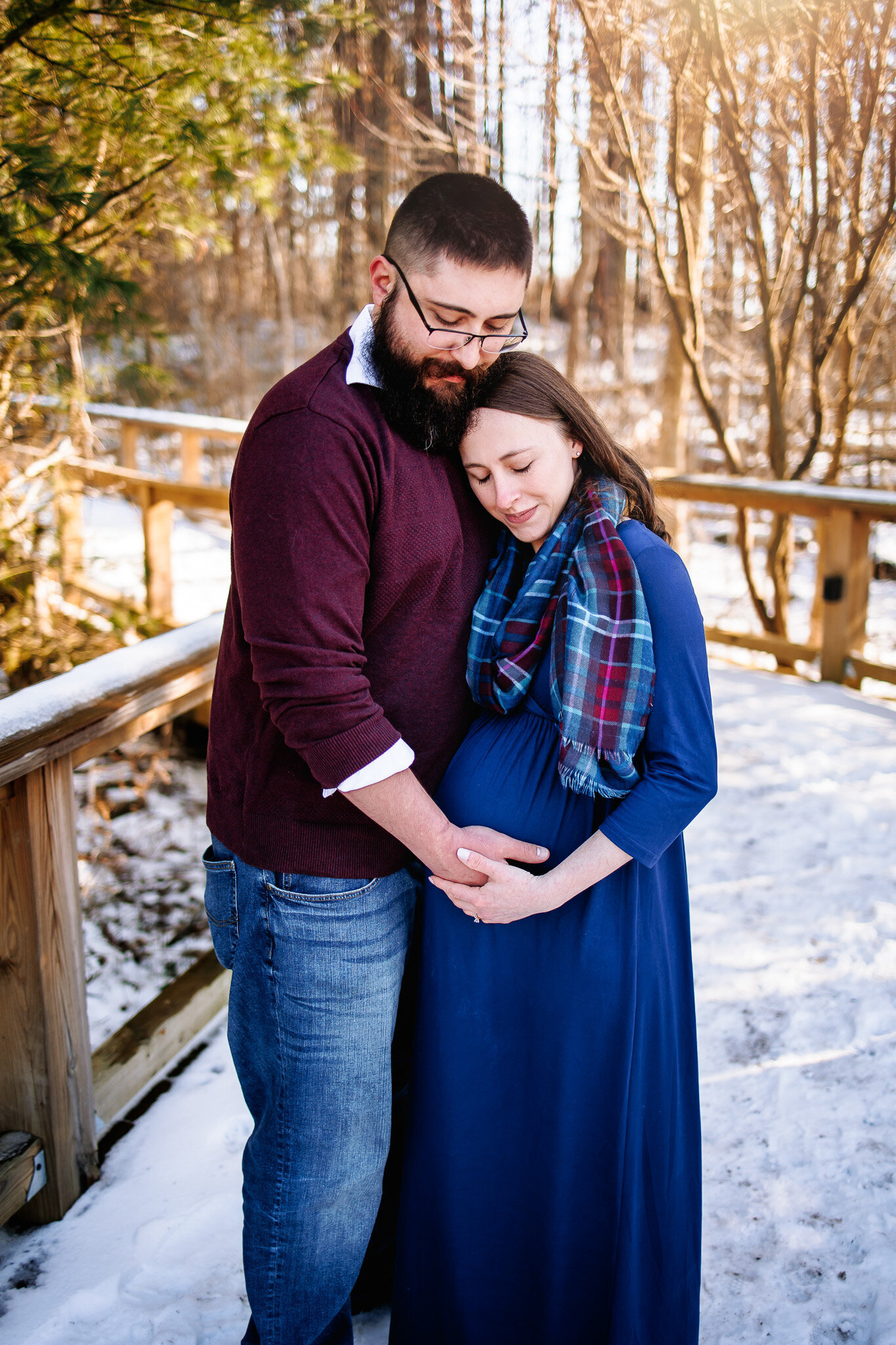 Expecting couple snuggle together while holding their baby to be amongst a snowy wooden walkway 