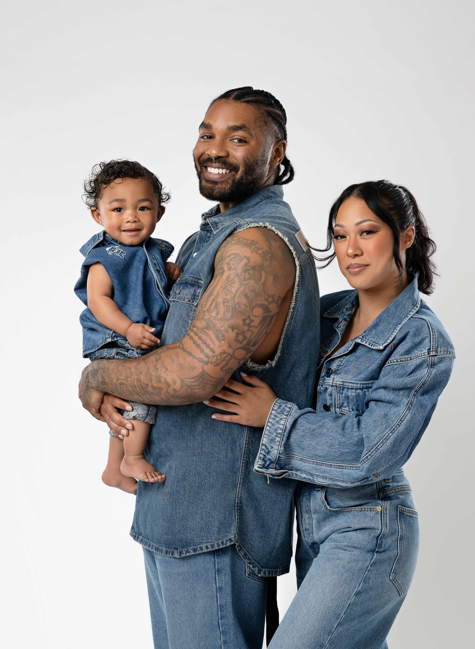 Family photo in jeans on white backdrop. Dad is holding baby and Mom is hugging in behind Dad.
