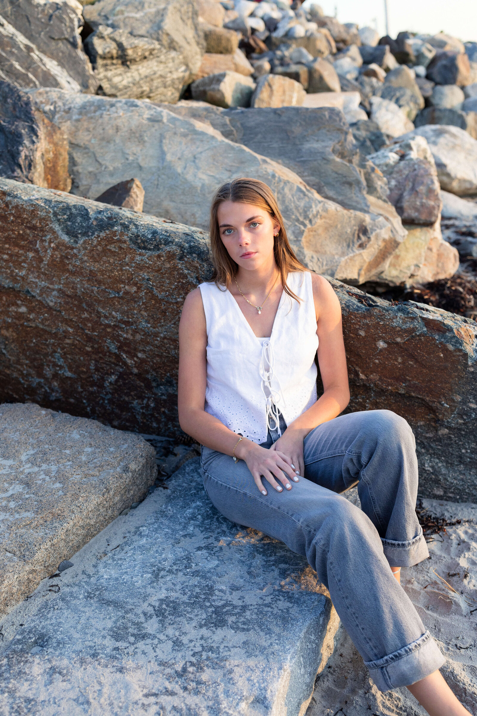 brunette high schooler posing on a beach wearing a white shirt and blue jeans on a rocky beach