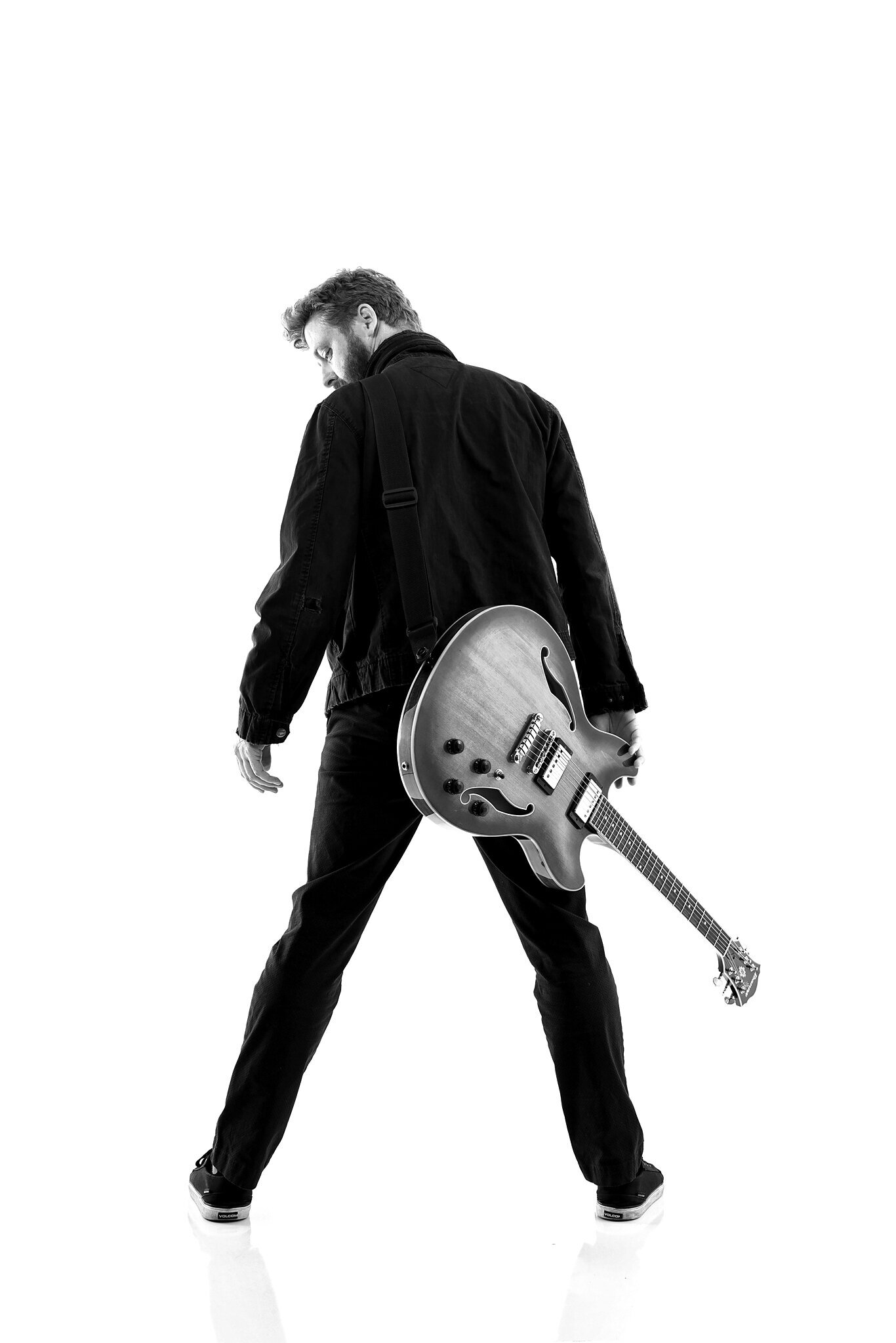 A man in dark clothing stands with his back to the camera, legs apart, holding an electric guitar by his side against a white background—capturing the essence of professional band photos Jacksonville musicians seek.