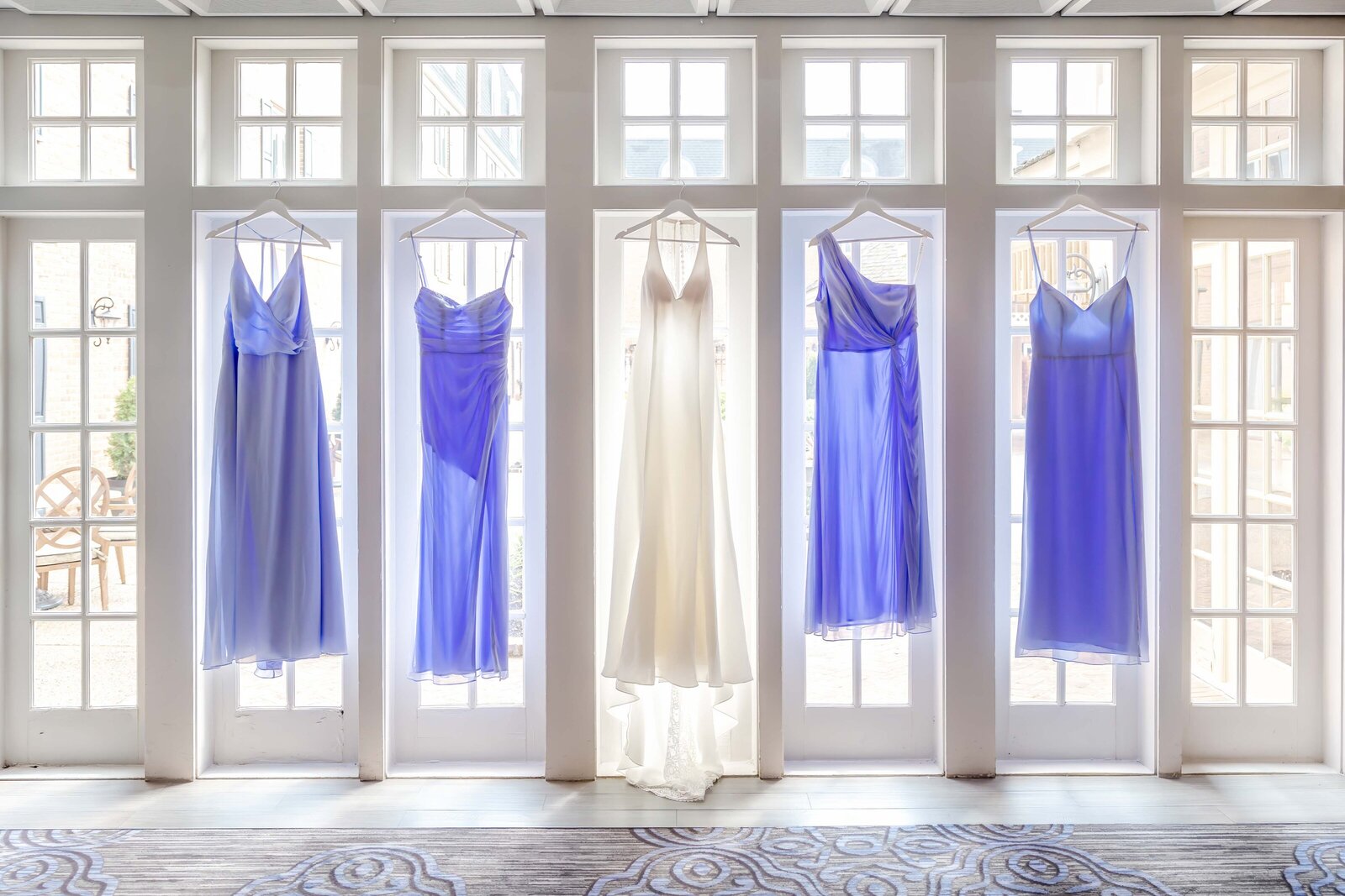 A wedding dress surrounded by lavender bridesmaid dresses displayed on hangers in front of large windows with natural light, captured during a Wilmington, PA wedding, showcasing elegant bridal preparations.
