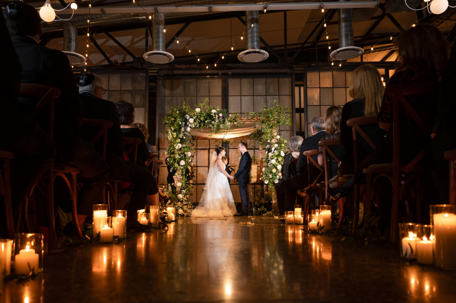 A couple stands under a floral arch during a wedding ceremony, with guests seated on either side and candles lining the aisle.