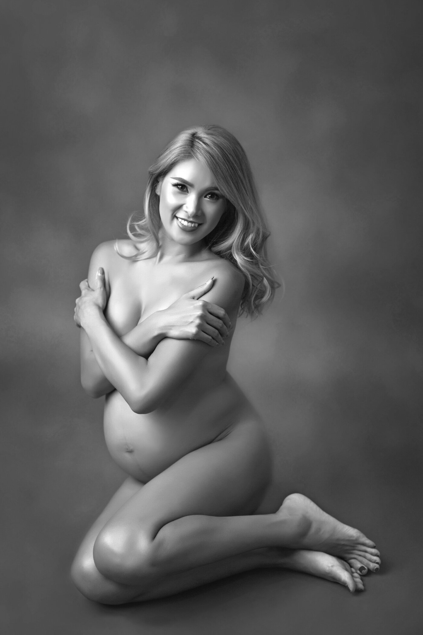 A black-and-white portrait of a pregnant woman kneeling on the ground, posing nude with her arms crossed over her chest to cover herself modestly. She smiles warmly at the camera, her long hair cascading over her shoulder. The soft lighting and subtle shadows highlight the natural beauty and grace of her pregnancy, creating an intimate and empowering image.