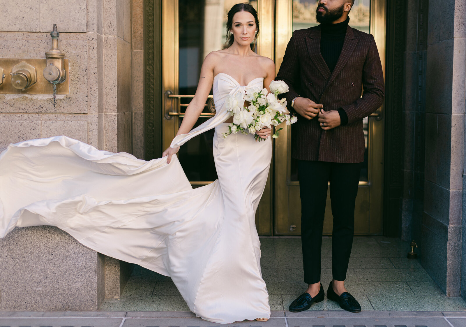 Editorial wedding portrait of a modern bride and groom in an urban setting, featuring a flowing silk gown and white bouquet with a fashion-forward twist.