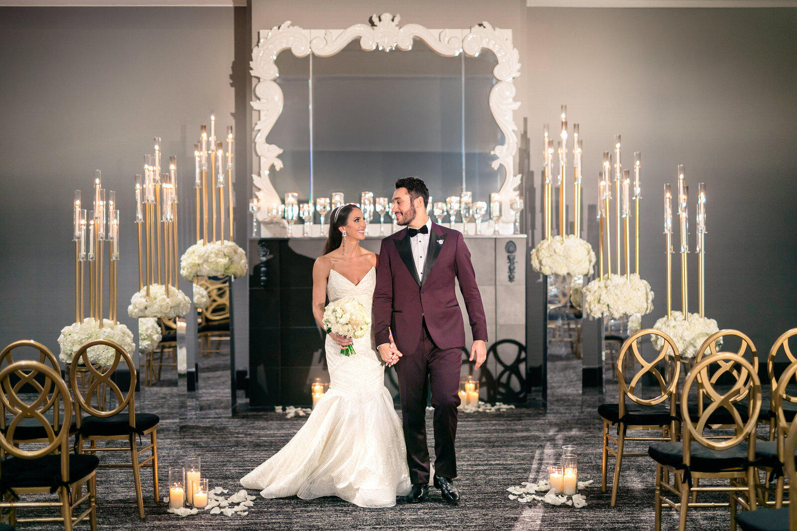 A bride in a white gown and a groom in a maroon tuxedo hold hands, smiling as they walk down an elegant aisle adorned with tall candles, white flowers, and decorative chairs. A large ornate mirror frames the background.
