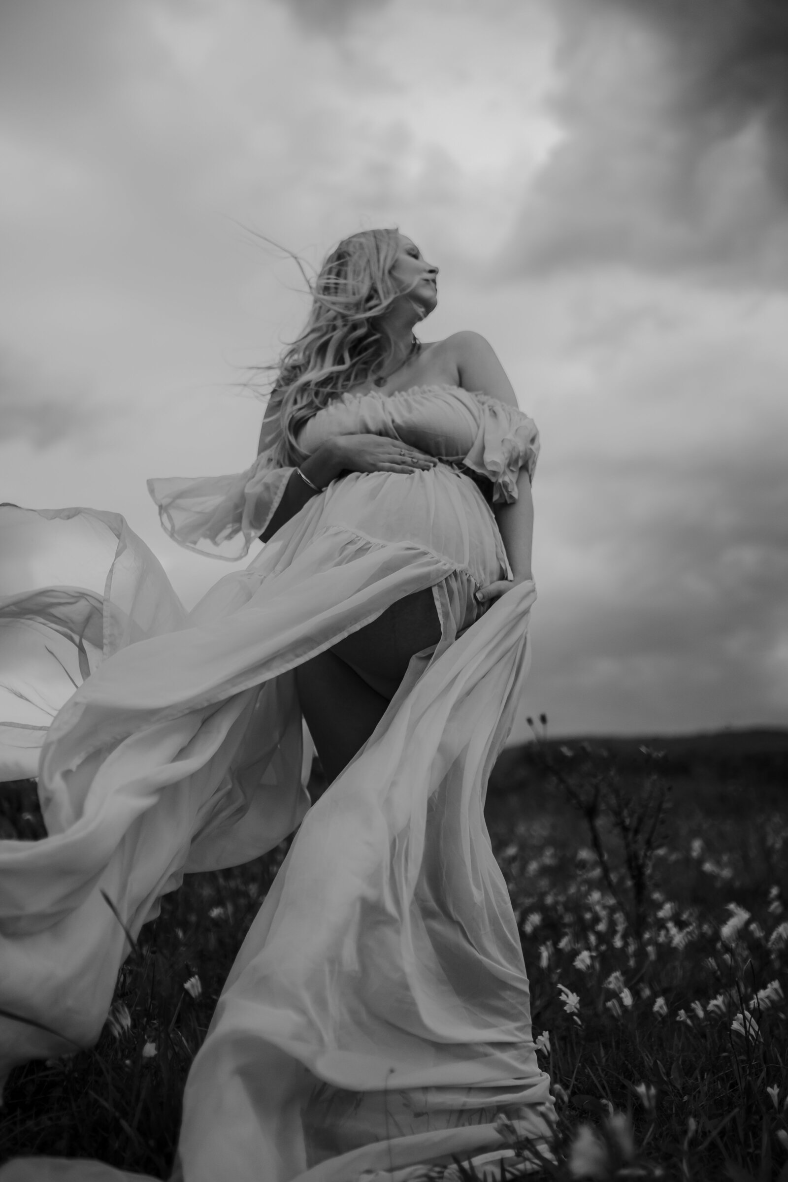 Black and white photo of a woman in a flowing dress standing in a field, looking upwards. The dress and her hair are being blown by the wind, and dark clouds loom in the sky above, evoking a sense of motherhood as she embraces the journey ahead.
