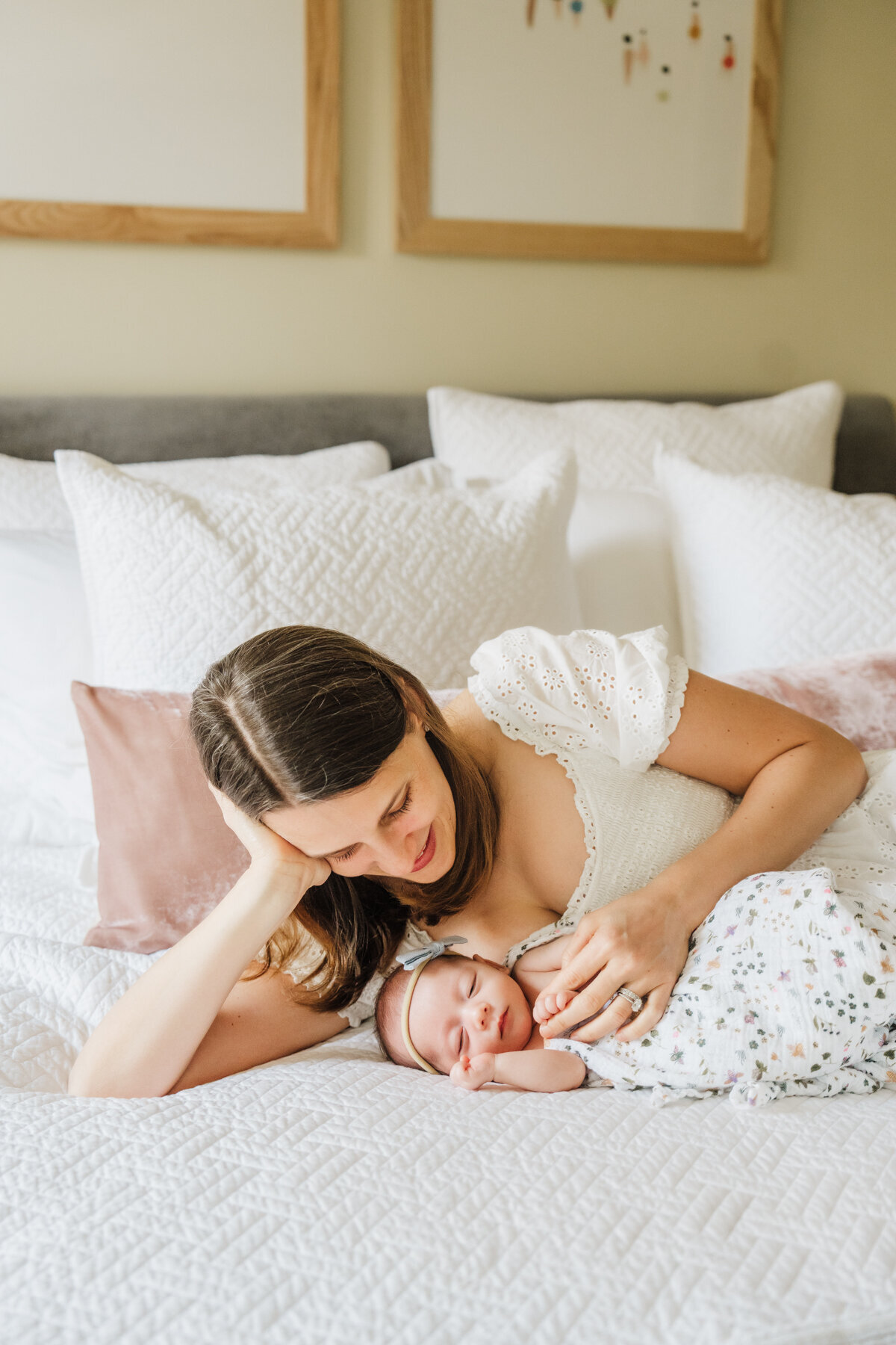 mother in white dress lays on white bed with pink pillows and admires infant daughter