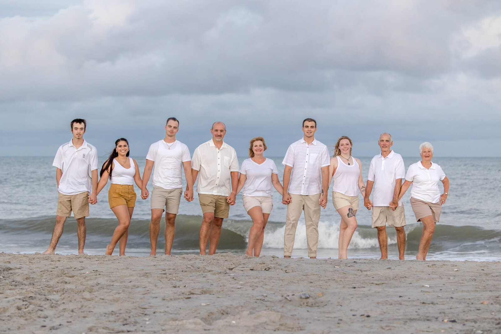 A large family stands hand in hand on the beach under a dramatic cloudy sky, dressed in white and khaki outfits. The ocean waves add a dynamic backdrop, perfect for North Carolina family photography, capturing unity and elegance.