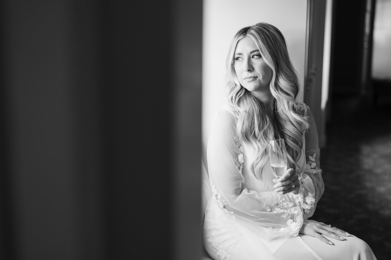 Bride holds a glass of champagne and looks out the window while getting ready