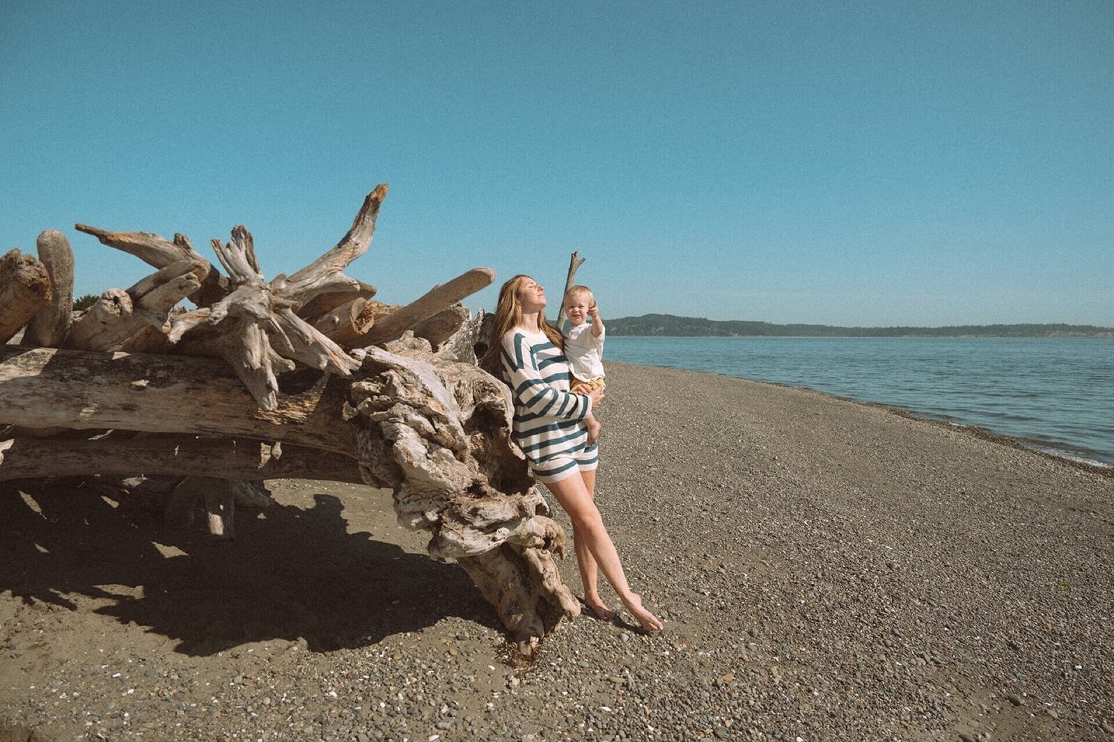 mother leaning on driftwood wearing blue stripes with little boy at beach on sunny day
