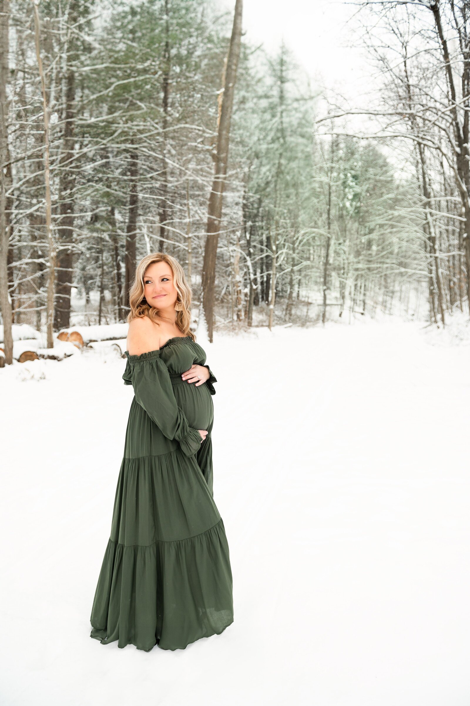 woman holding her pregnant belly in a green dress while standing on a trail covered in snow.