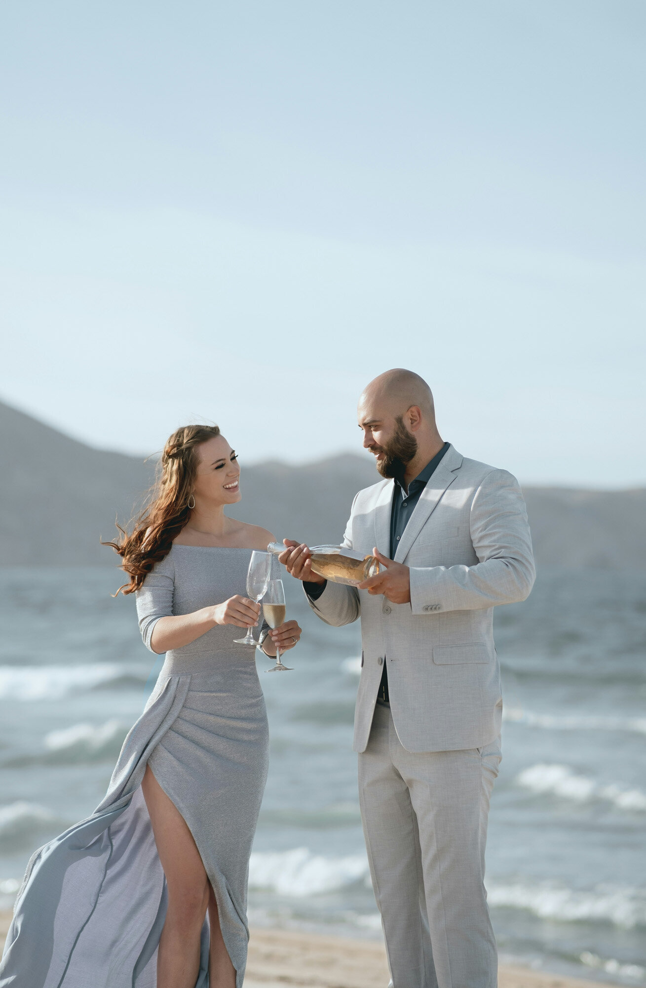 Couple on the seashore while woman holding two wine glasses and man holding a bottle of wine pouring it to the wine glass