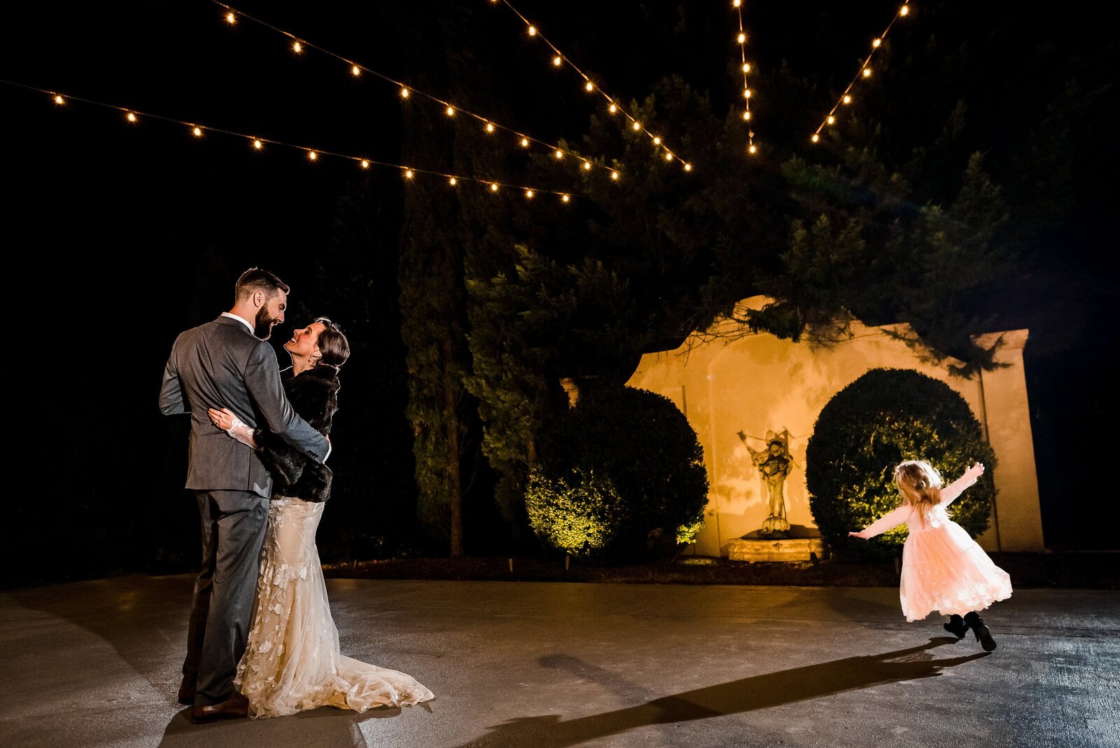 flower girl runs onto the dance floor while the bride and groom share their first dance on an outdoor patio under string lights