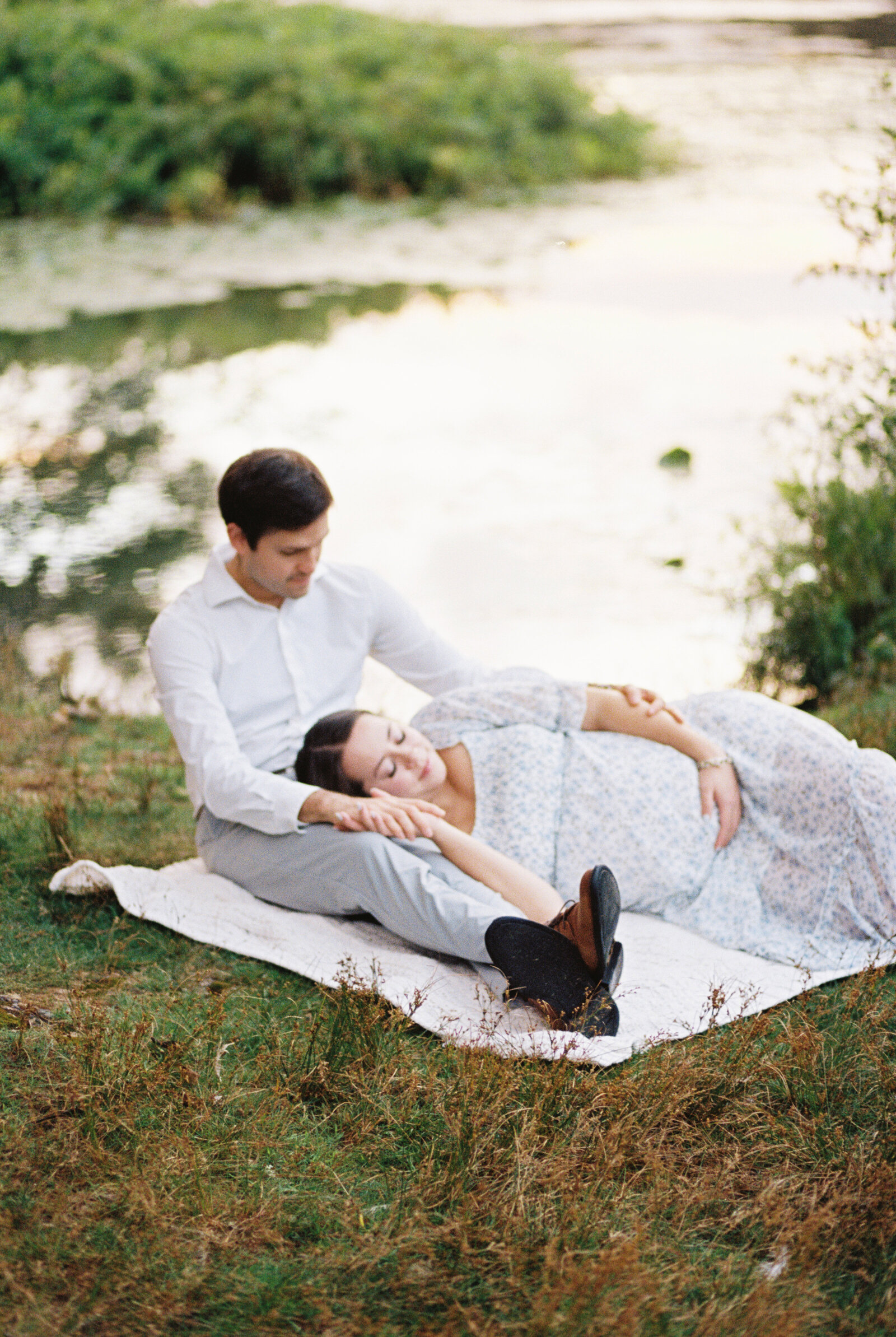 A pregnant woman is laying down on her husbands leg while he sits on a blanket