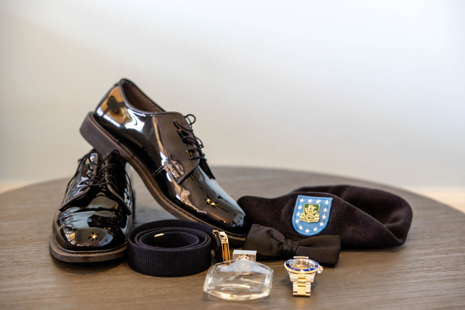 A flat lay of groom’s polished black shoes, bow tie, belt, cologne, watch, and military beret, captured at a Carolina Beach, NC wedding to highlight elegant and meaningful wedding day details.