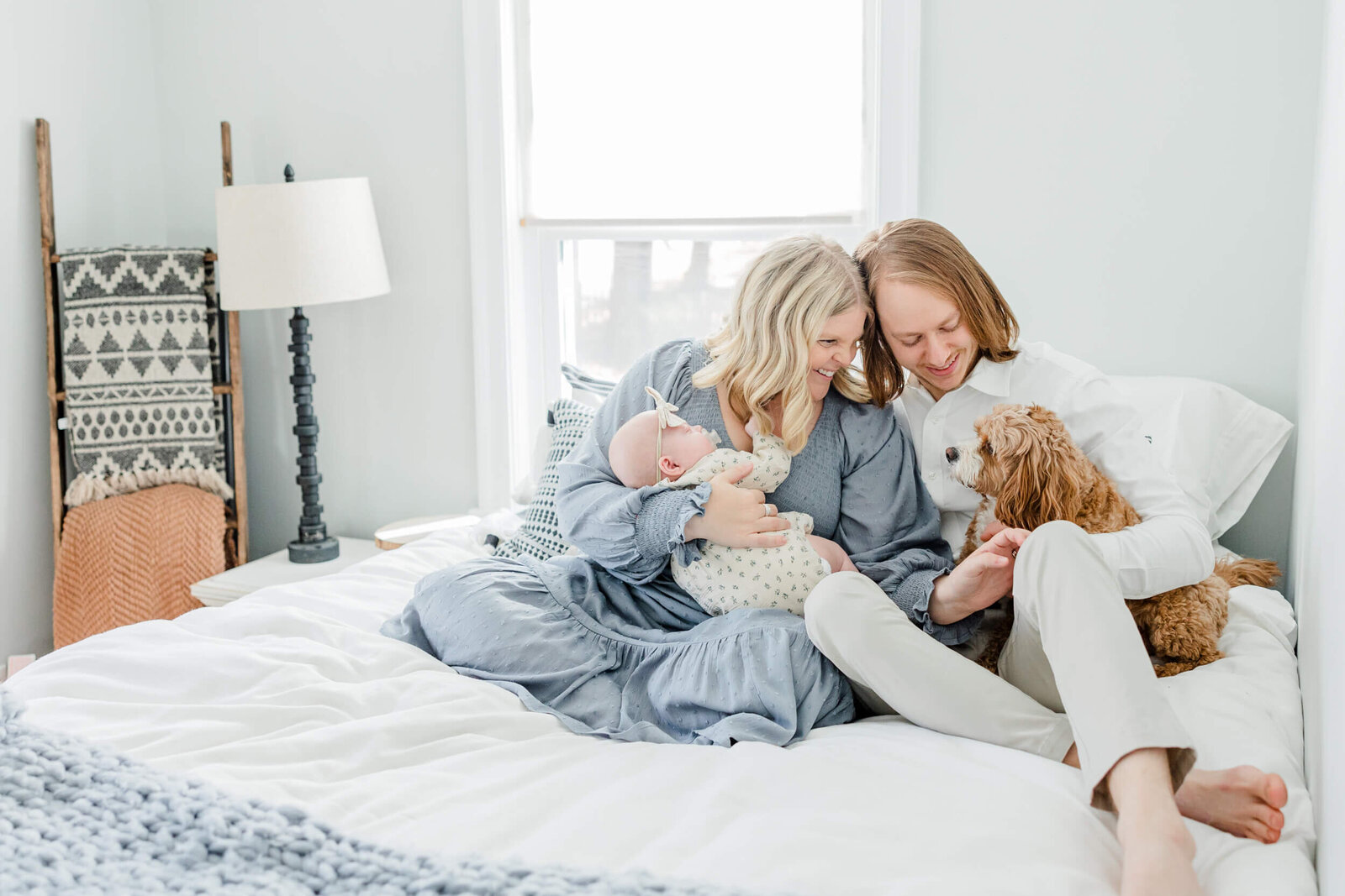 Mom holds newborn and sits with dad and their dog on a bed