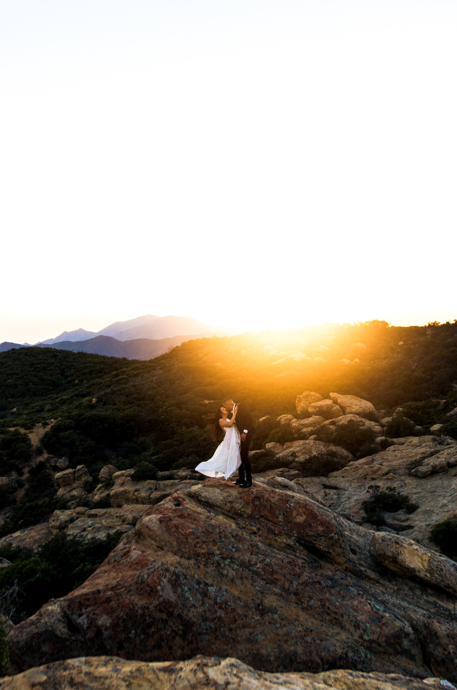 Wedding couple, standing on large rock kissing with mountains and sunset in the background.