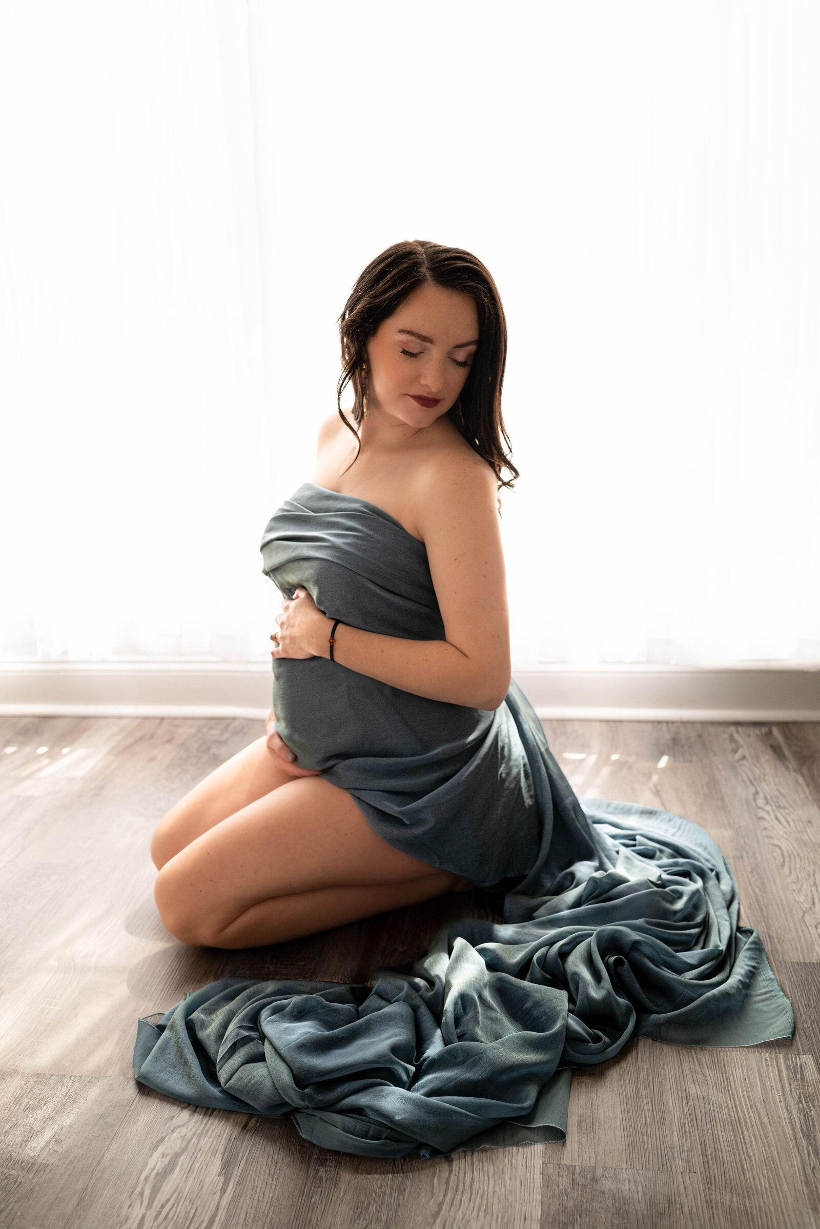 A pregnant woman draped in  blue fabric and kneeling on the floor for her studio maternity photos in Huntsville Alabama