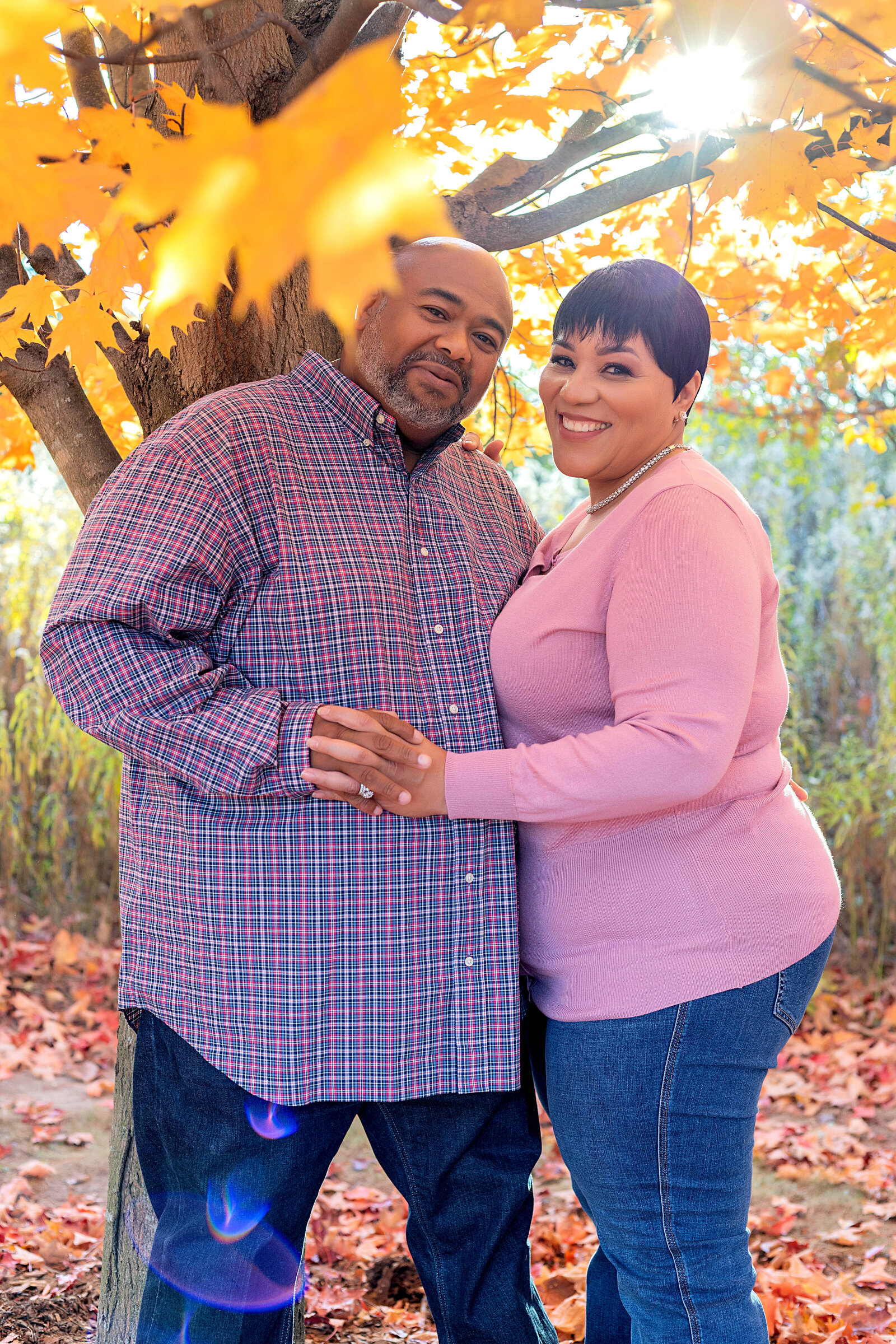 Married couple wearing blue and pink plaid top, pink sweater, and blue jeans standing under trees