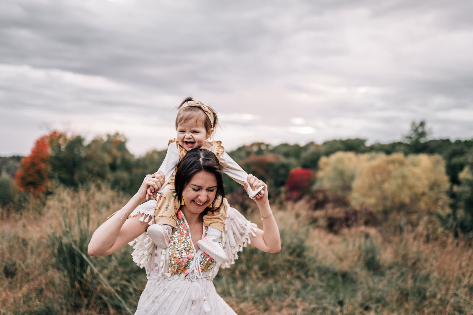 A mother, dressed in a white, embroidered dress, holds her young child on her shoulders as they walk through a grassy field.