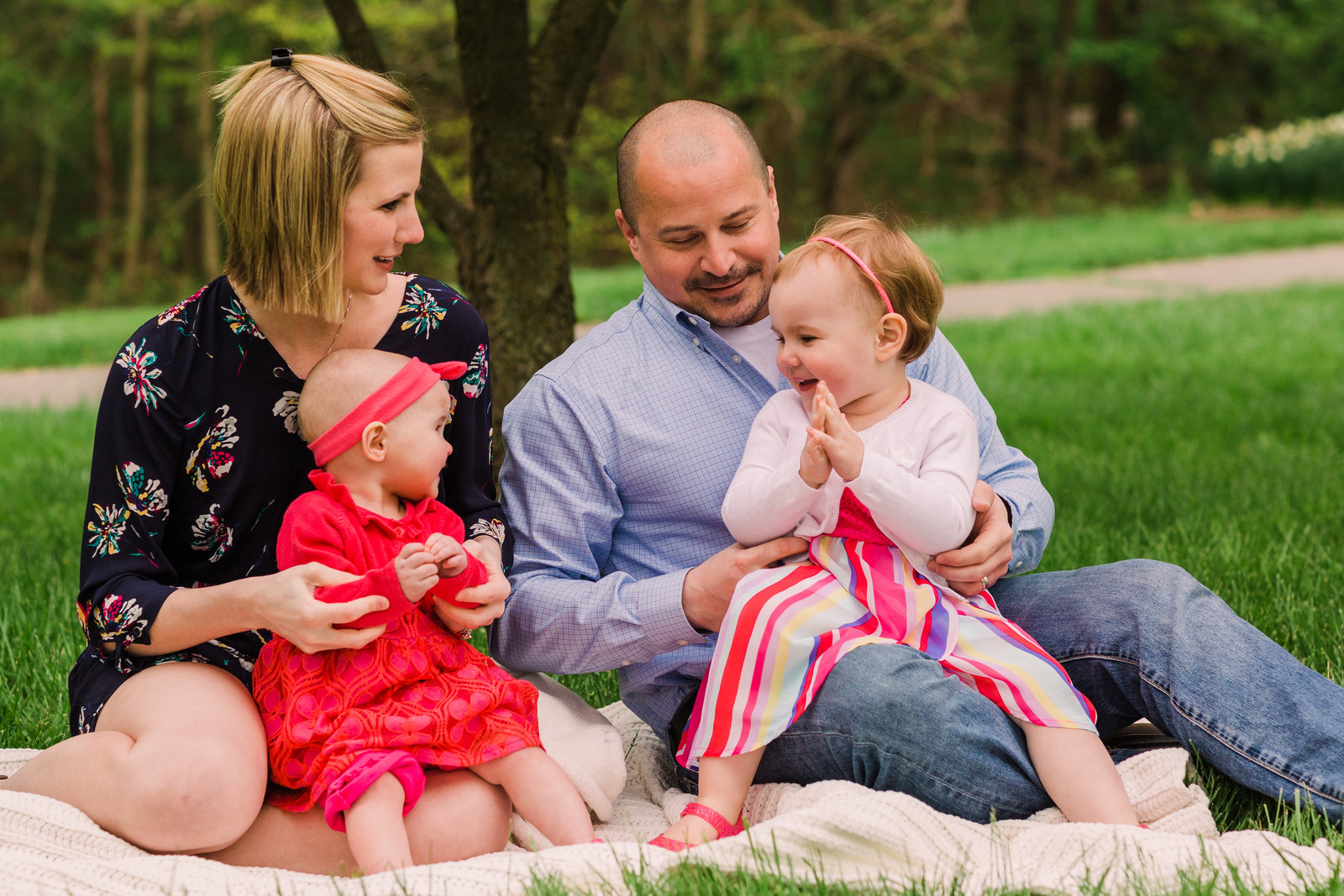 A family of four, mom, dad, and two young daughters pose for a photo  sitting on lush green grass