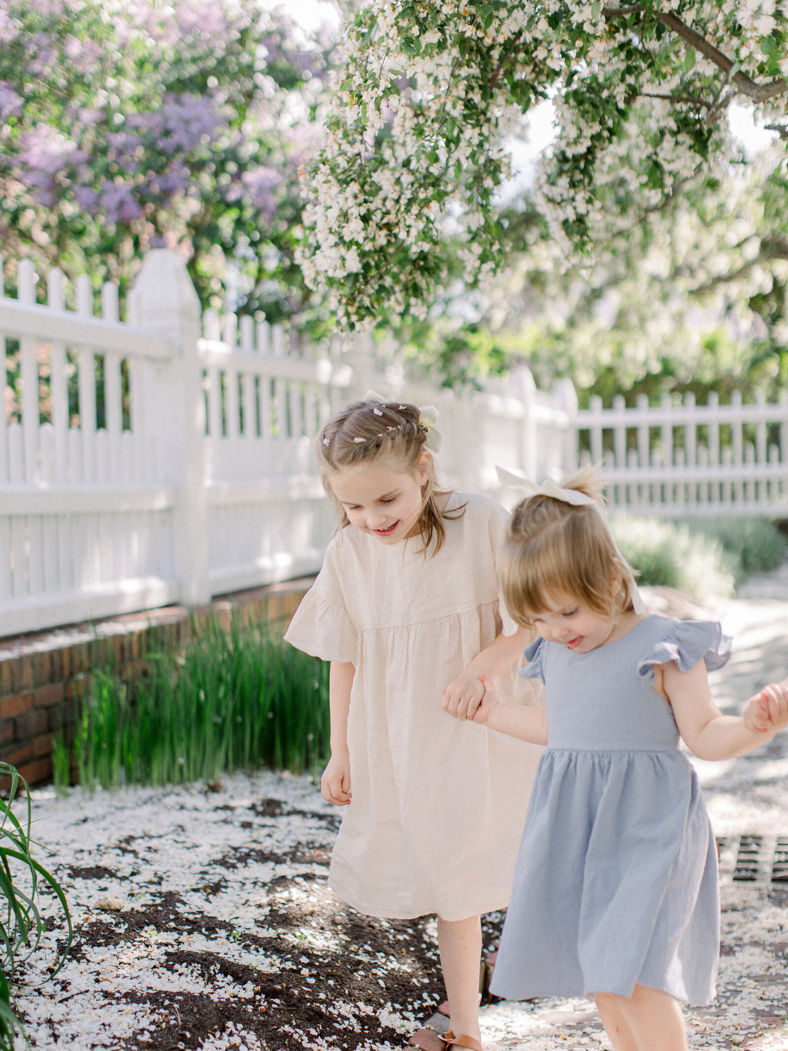 Sisters in linen dresses holding hands and walking through a garden of flowering cherry trees by NH newborn photographer Fieldstone Studio.