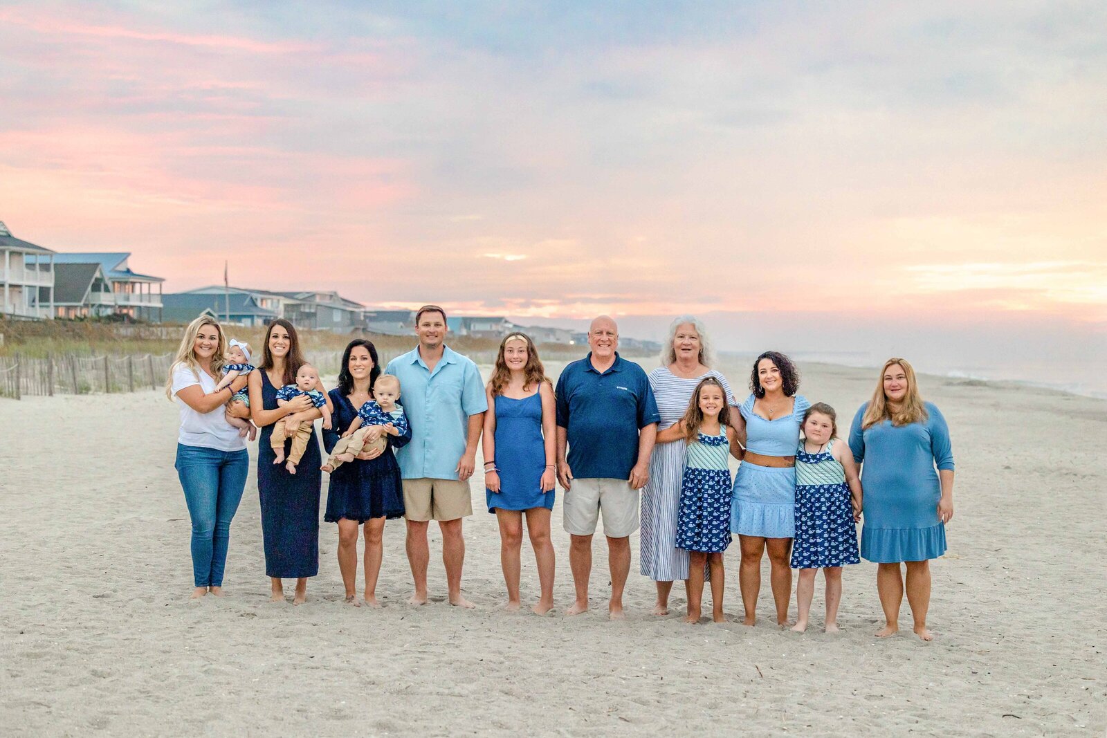 A large family stands together on Ocean Isle Beach at sunset, dressed in blue and white attire. The colorful sky and beachfront homes add warmth to the scene. Ideal for artistic family photography in Wilmington, NC, capturing vibrant family moments.