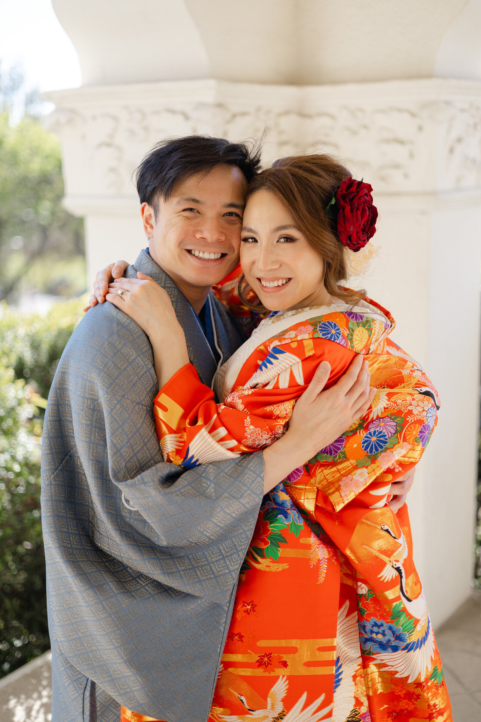 Bride in red iro uchikake and groom in formal montsuki embracing at Brand Park Glendale, Los Angeles California. Available for romantic wedding kimono rental | 赤い色打掛の花嫁と紋付袴の新郎がブランドパーク・グレンデールで抱き合うロマンチックなひととき。ロサンゼルス カリフォルニアの着物レンタル。