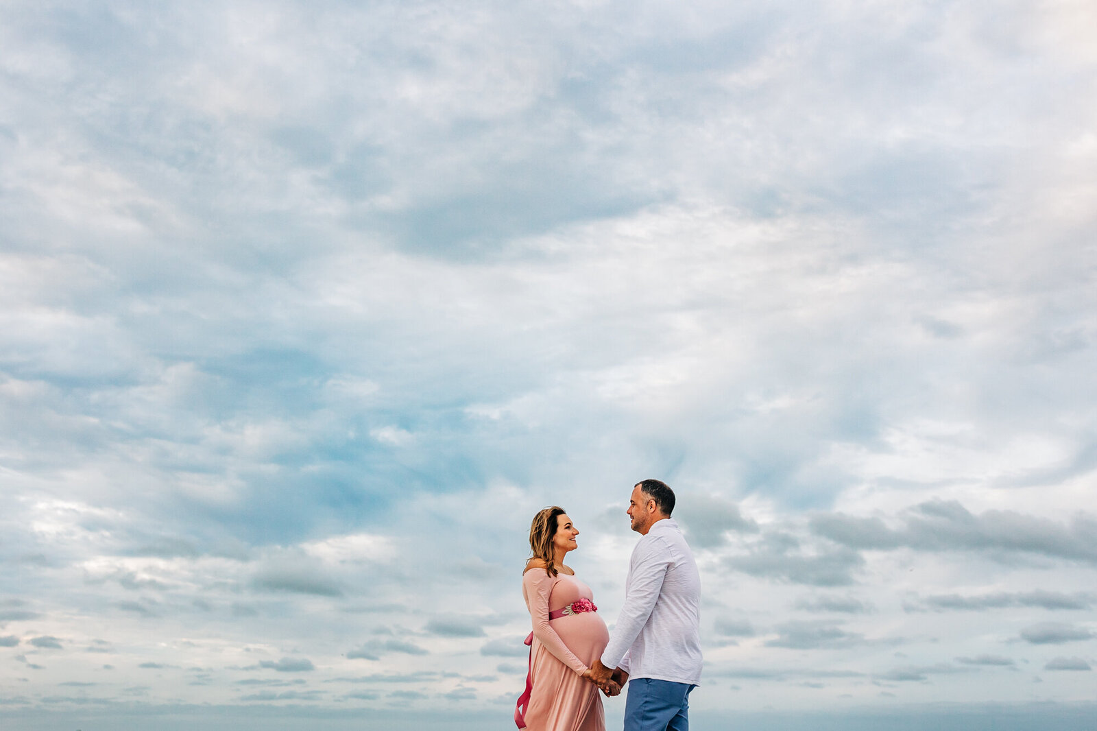 An expecting couple embracing on the beach with a cloudy sky behind them