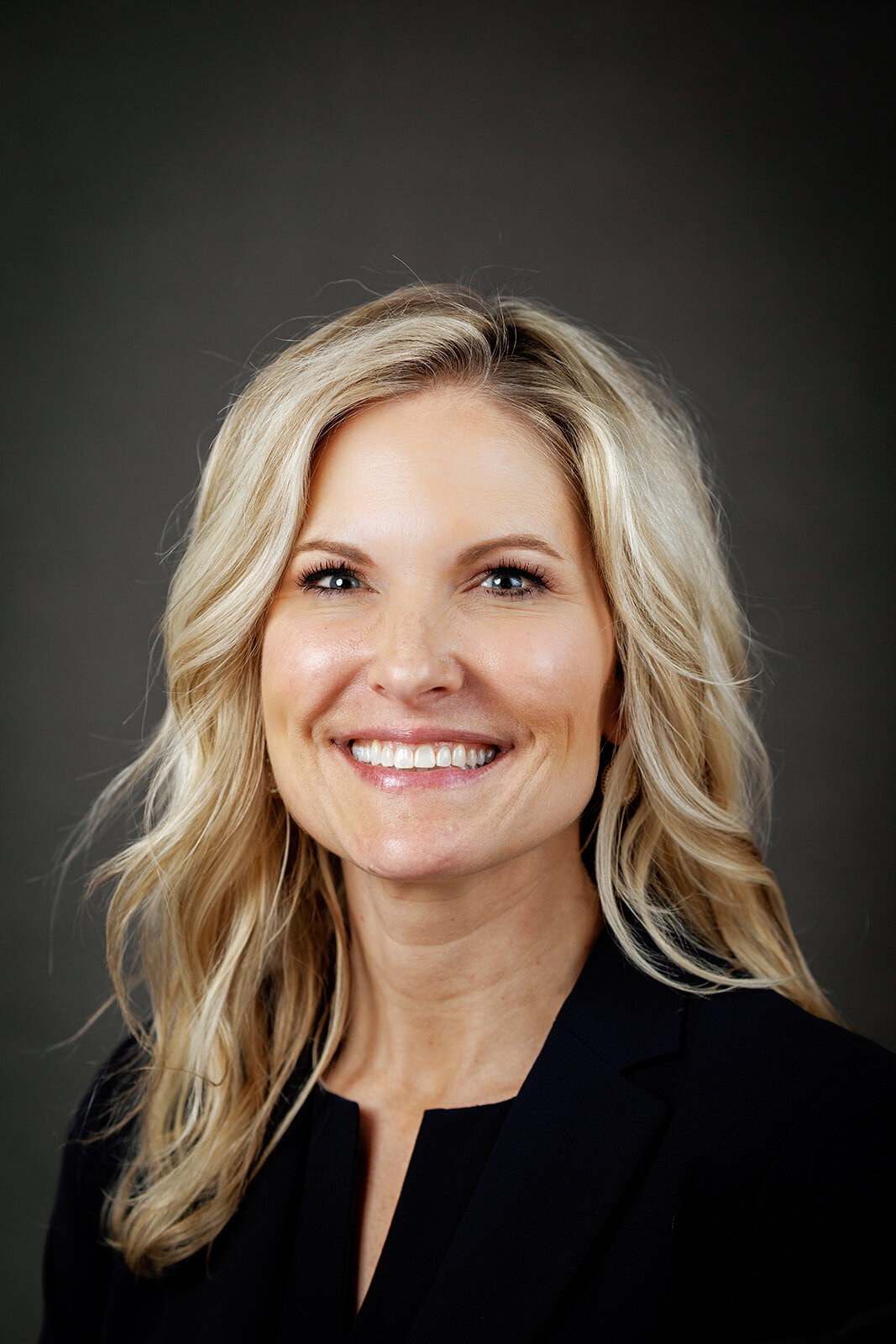 Corporate headshot of a woman with blonde hair and a black blazer, smiling brightly against a neutral dark background.