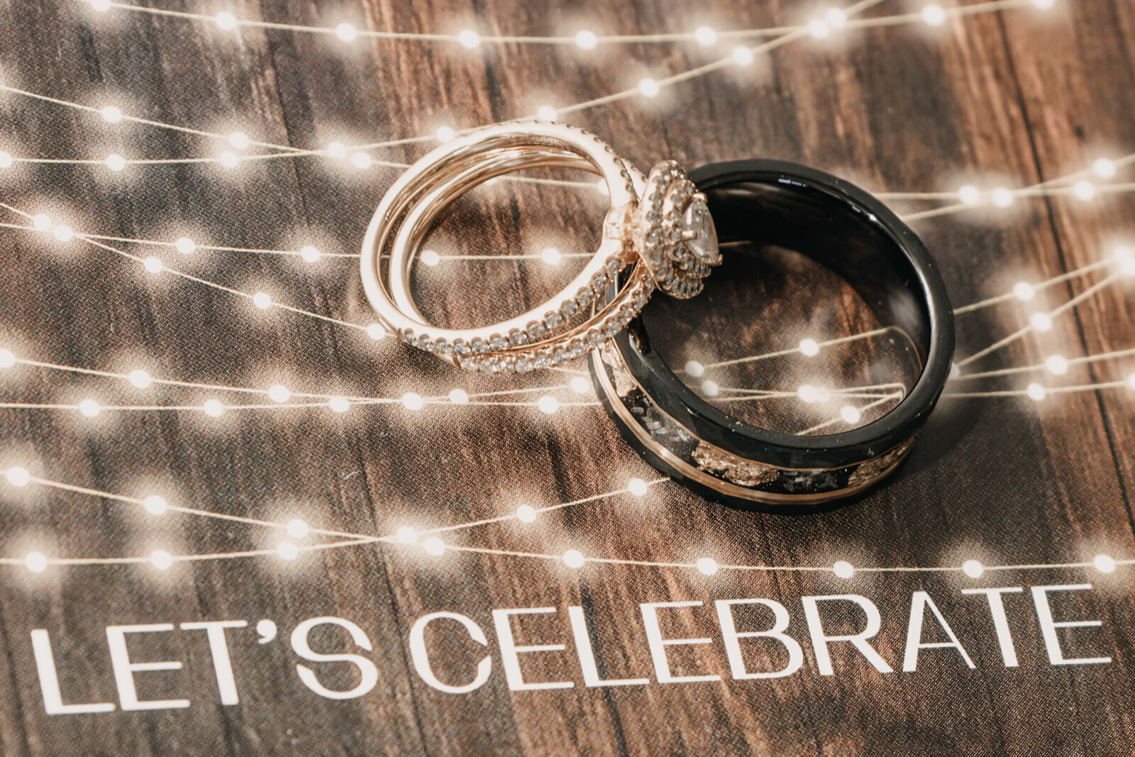 A pair of wedding rings resting on a wooden table, surrounded by the warm glow of string lights above.
