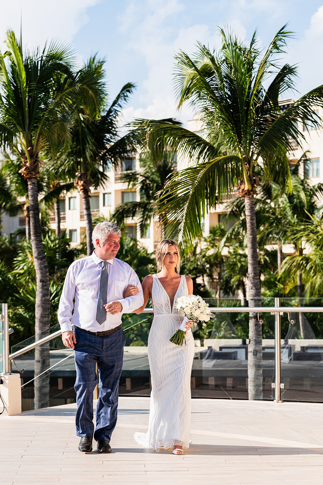 A beautiful moment as the bride walks arm in arm with her father down the aisle towards her wedding ceremony, framed by palm trees and the ocean in the background. Captured by CMH Studios during a destination wedding