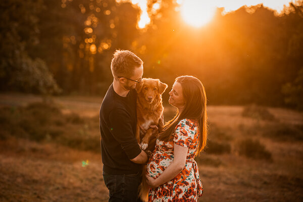 Een aanstaande moeder en haar partner poseren liefdevol met hun hond tijdens een zwangerschapsshoot, een intiem moment vol warmte en verwachting