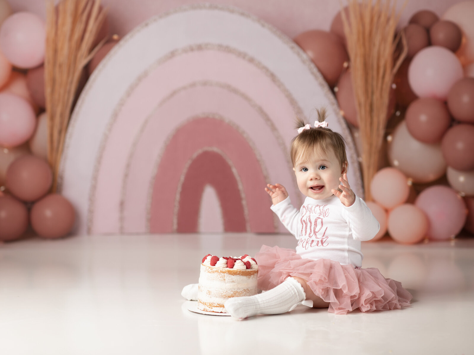 one year old girl sitting with cake and boho rainbow backdrop for cake smash photoshoot