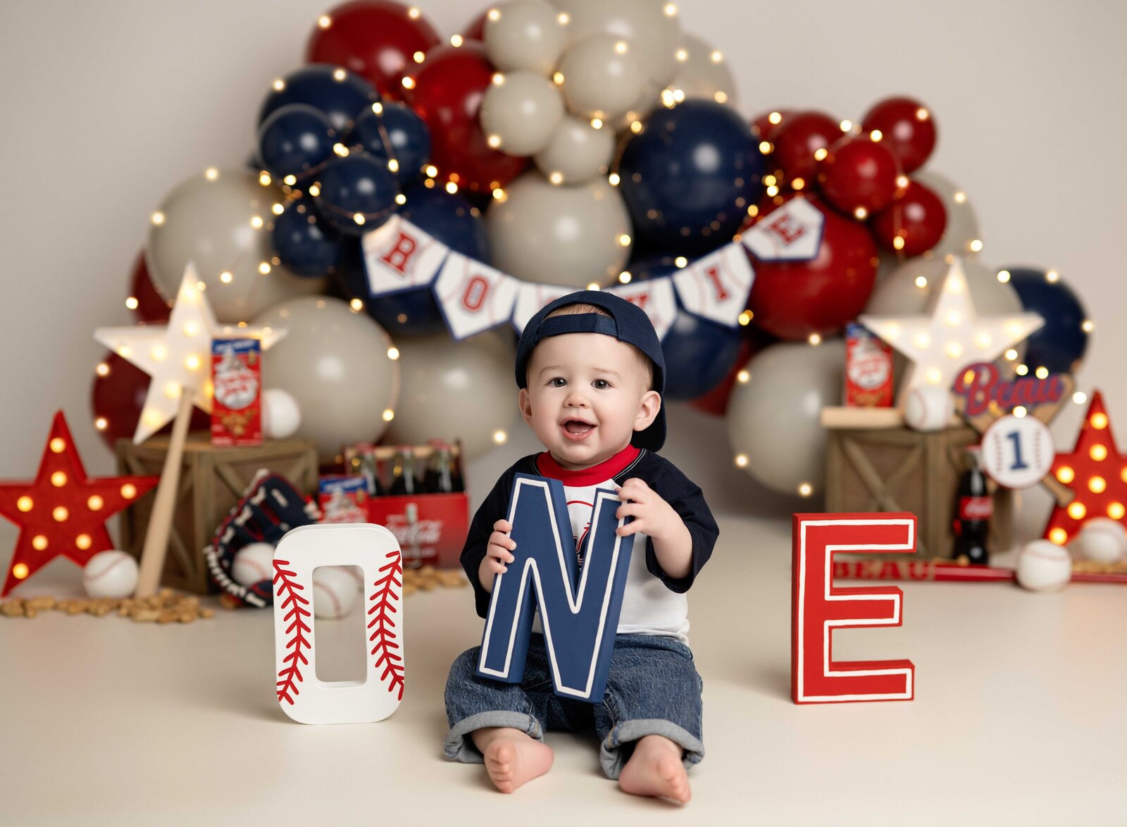 Baby boy in baseball shirt holding baseball theme ONE letters and smiling. Studio cake smash photo taken by Jenni Nikki Photography in Atlanta, GA.
