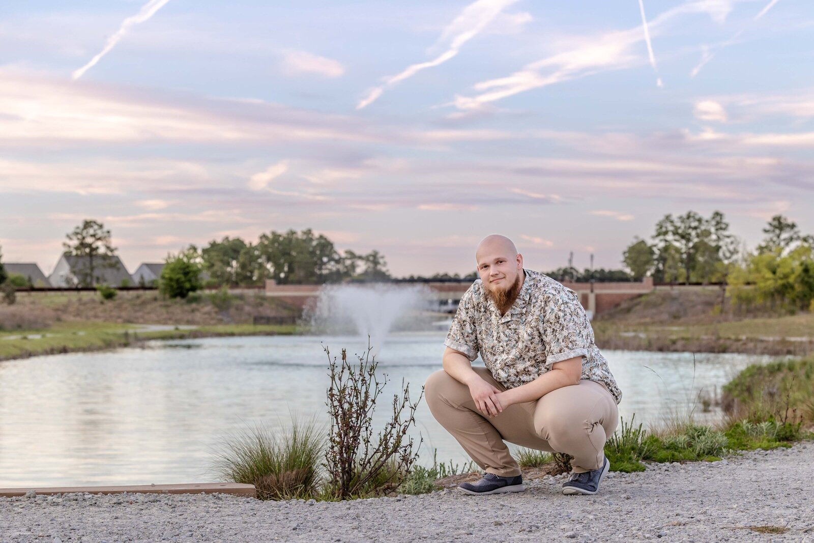 A college senior crouching by a lake with a fountain backdrop during a colorful sunset in Wilmington, NC. This relaxed portrait showcases professional senior photography, perfect for capturing natural and serene moments.