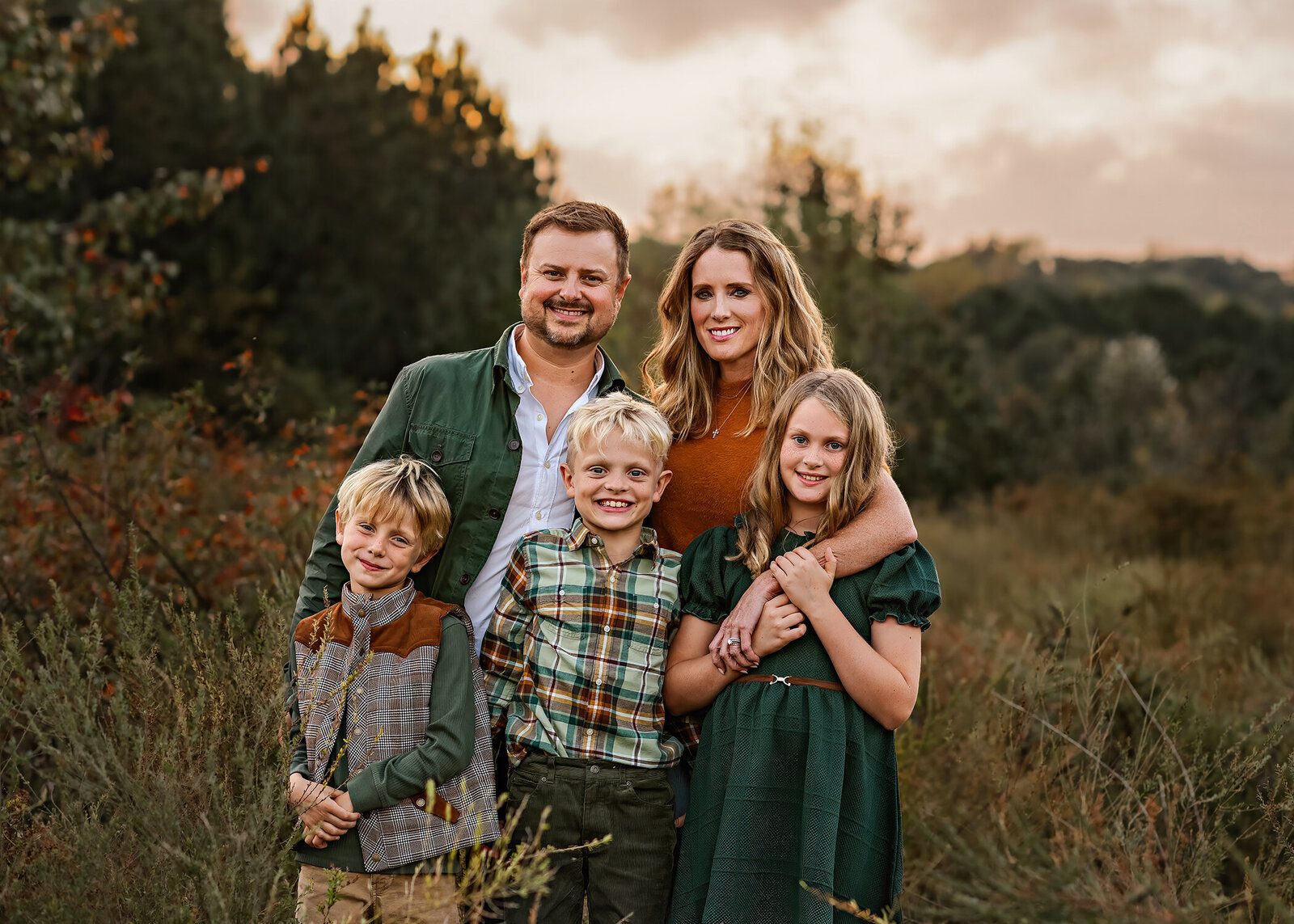 A family of five stands close together in a field at golden hour, with a soft, cloudy sky and trees in the background. The father, wearing a green jacket, and mother, in a rust-colored top, smile as they wrap their arms around their three kids in front—two blond boys in plaid and a green vest, and a girl in a dark green dress. Everyone is looking at the camera with warm, relaxed expressions.