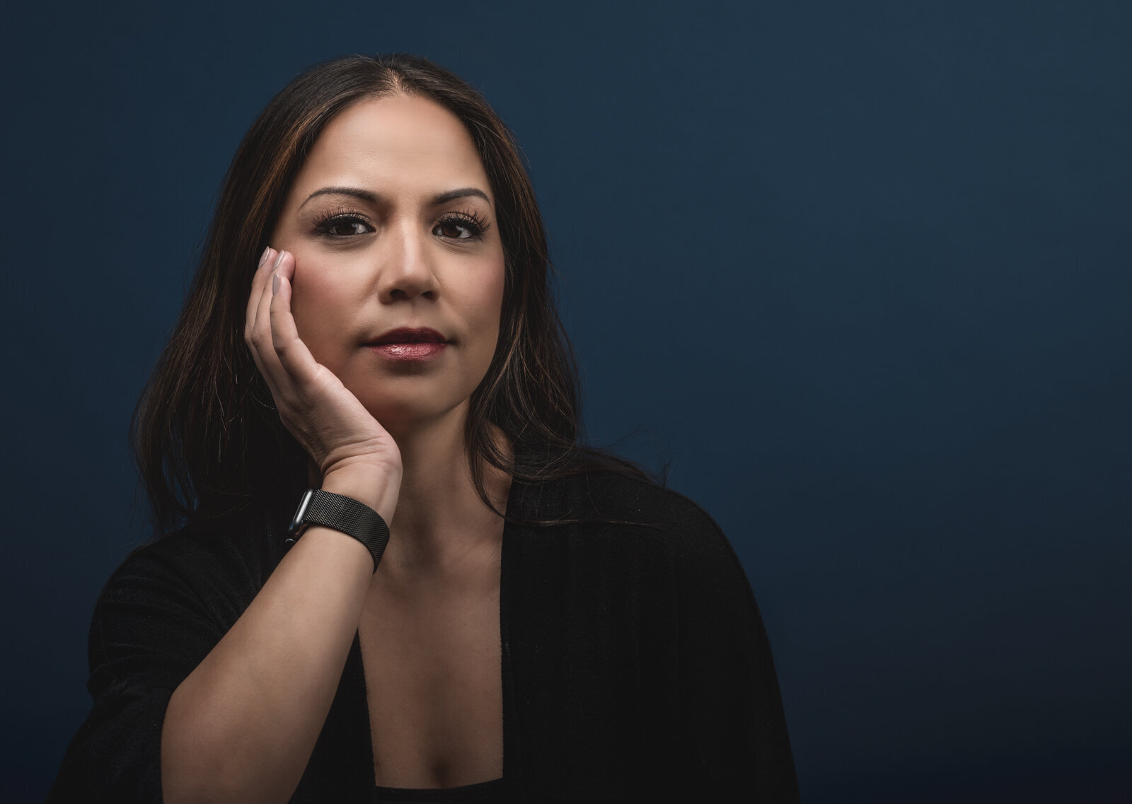 Woman sits with her right hand resting on her face wearing a black top against a navy background for a headshot session in Oakland, CA