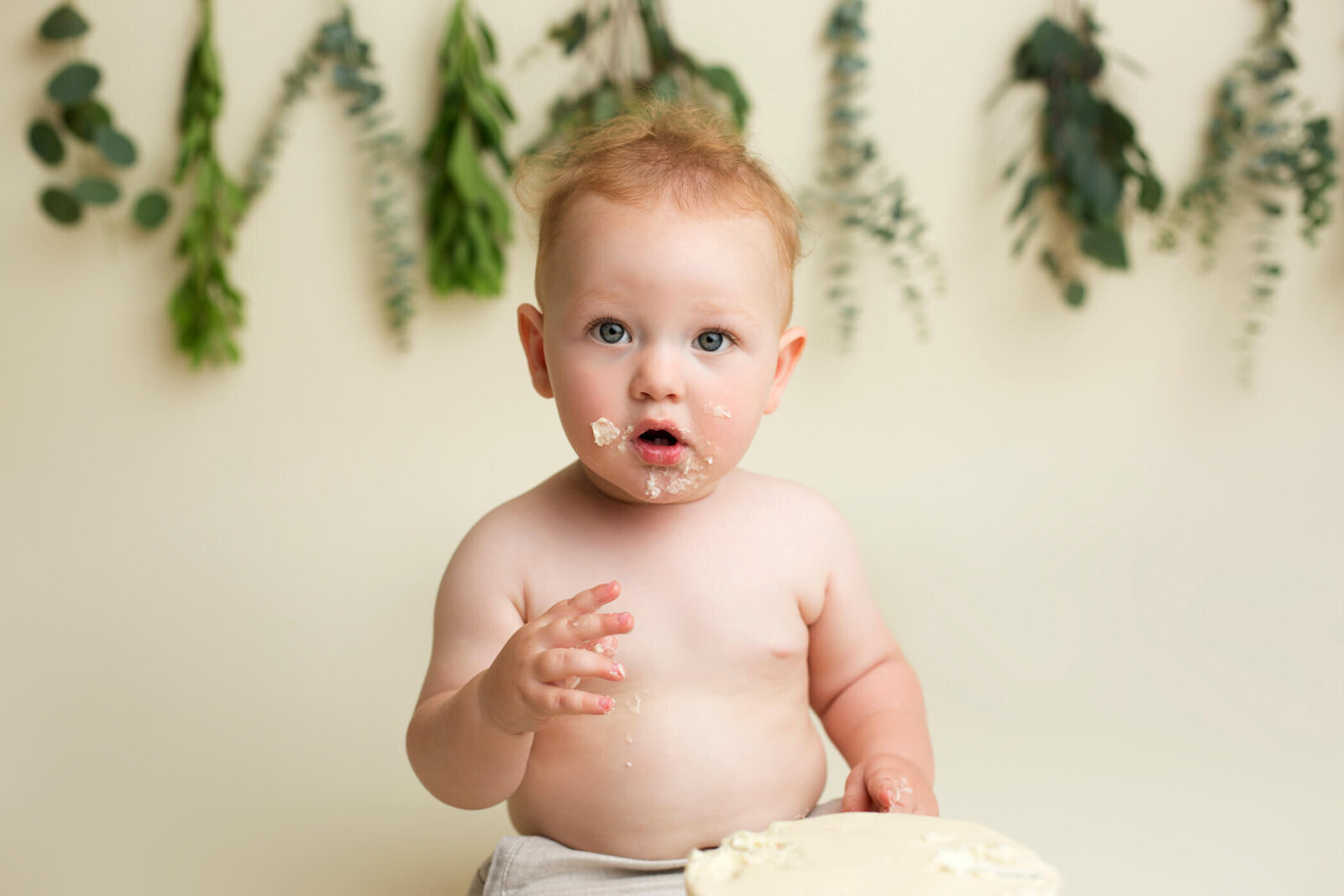 Adorable baby with short blond hair, sitting and eating a cake with frosting on their face and hand. The background features hanging greenery, creating a natural and festive setting.