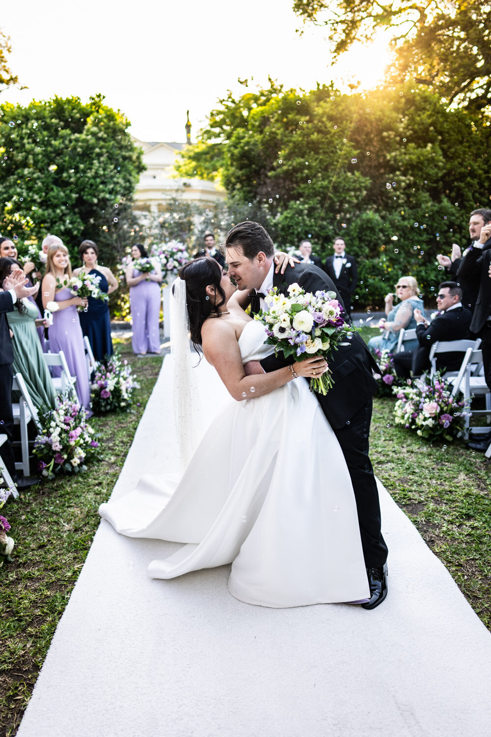 Bride and groom kiss while walking out at end of wedding ceremony at Elms Mansion, with bubbles and sunset in background