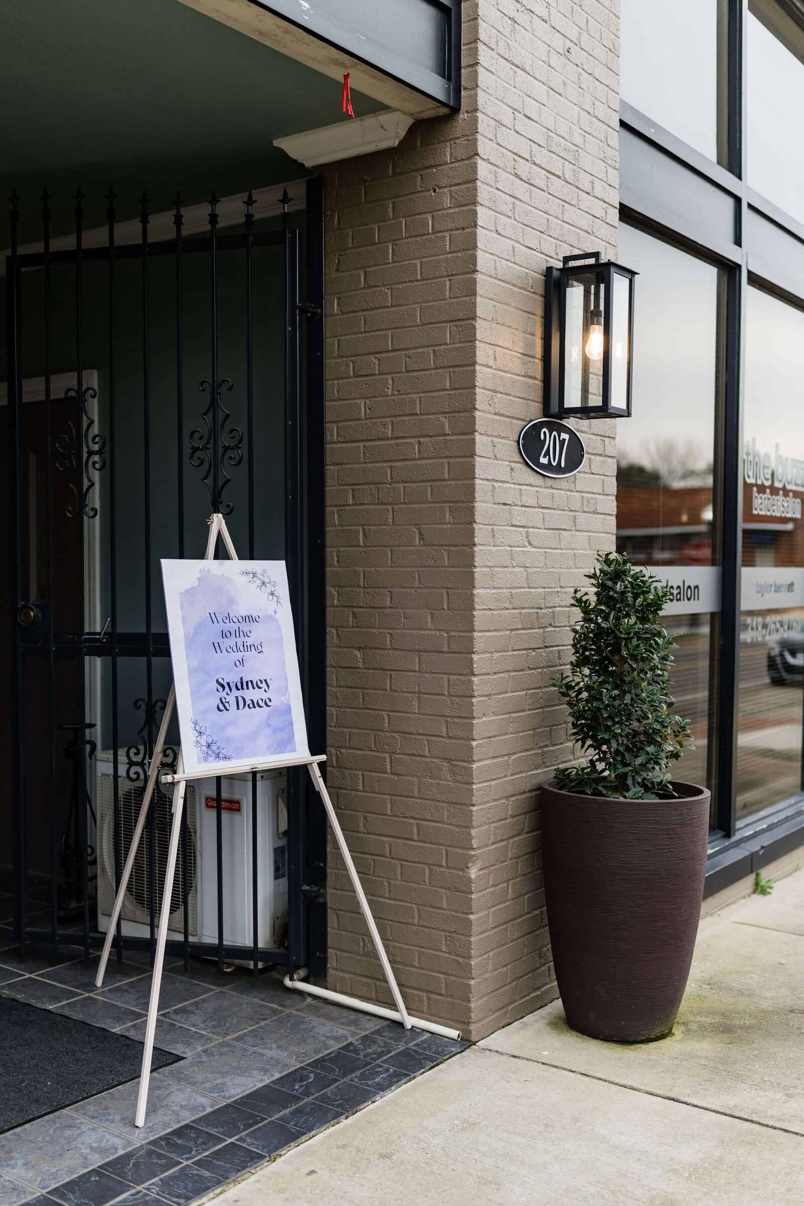 A sign on an easel near a the entrance of The Norton Building in Ruston, Louisiana reads Welcome to the wedding of Sydney and Dace. A potted plant and a lantern-style light fixture are beside the door, with the address number 207 on the wall.