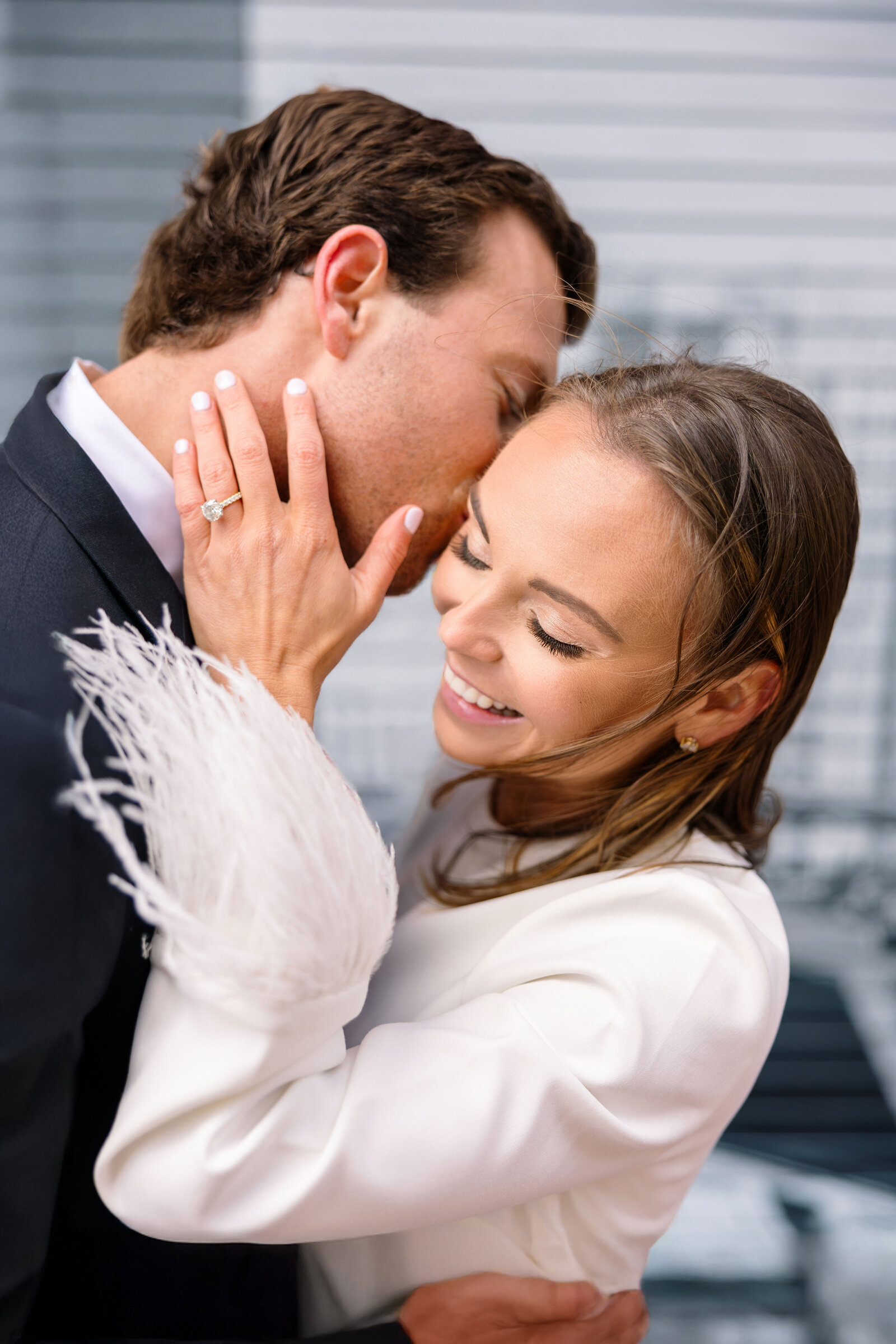 A couple embraces, smiling with joy. The man kisses the womans forehead as she displays an engagement ring, her hand resting on his cheek. They are dressed in elegant attire, with a cityscape blurred in the background.