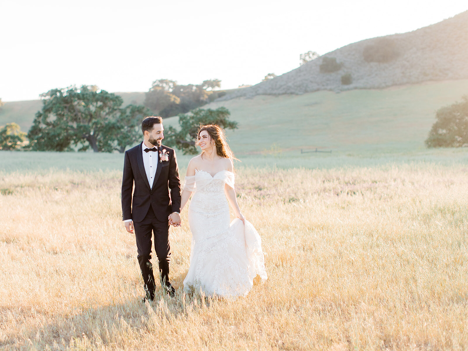 bride and groom holding hands and walking through a field at sunset